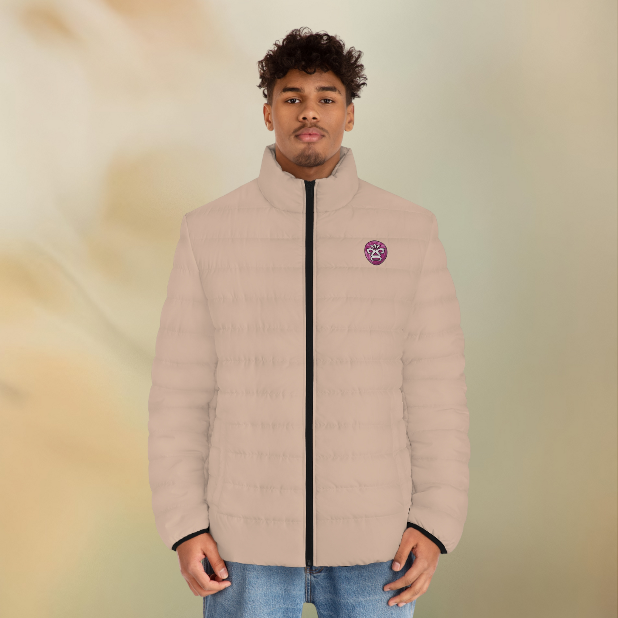 Ein männliclhes Modelt zeigt tragend die  Herren-Pufferjacke mit der Vorderansicht  in einem Wüstensand Farbton. Die Jacke ist horizontal gesteppt, hat eine glatte Oberfläche, einen hohen Stehkragen und einen durchgehenden Reißverschluss der Schwarz ist . Auf der linken Brusthöhe ist ein Emblem im Maori Stil. Designed, by Just a Girl

