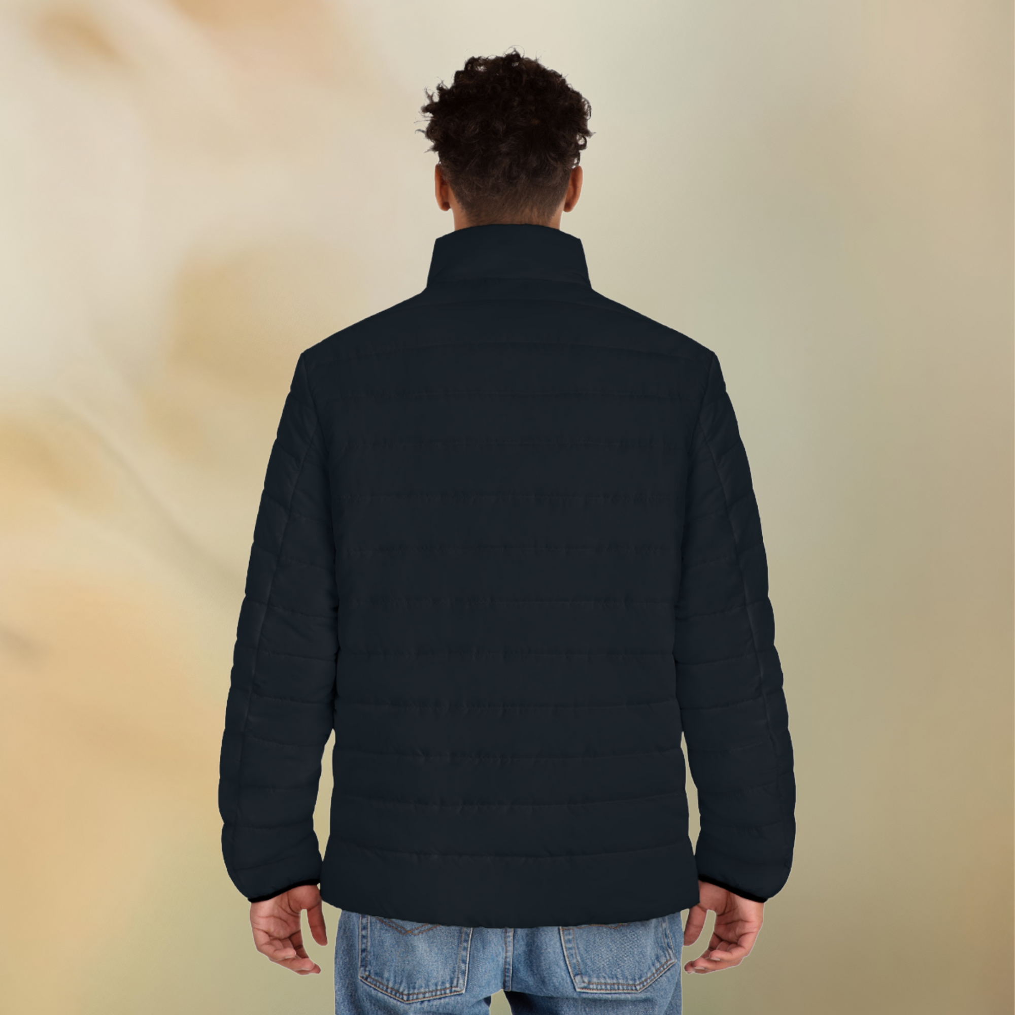 Ein männliches Model zeigt tragend mit der Rückenansicht die  Herren-Pufferjacke in einem natürlichen schwarzen Farbton. Die Jacke ist horizontal gesteppt, hat eine glatte Oberfläche sowie einen hohen Stehkragen.  Designed, by Just a Girl

