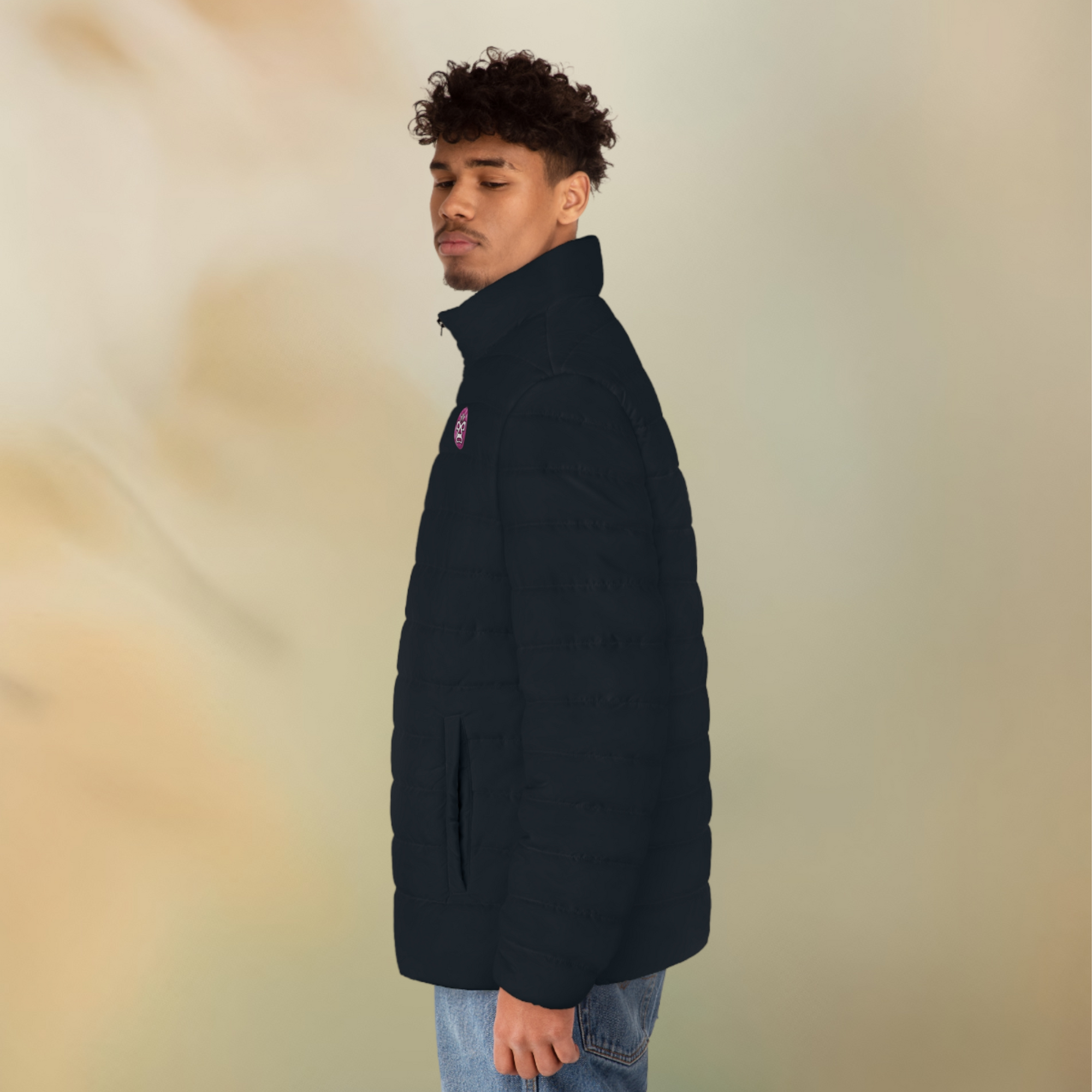 Ein männliches Model zeigt tragend von der linken Seite die Herren-Pufferjacke in einem natürlichen schwarzen Farbton. Die Jacke ist horizontal gesteppt, hat eine glatte Oberfläche, einen hohen Stehkragen und einen durchgehenden Reißverschluss der Schwarz ist . Auf der linken Brusthöhe ist ein Emblem im Maori Stil. Designed, by Just a Girl

