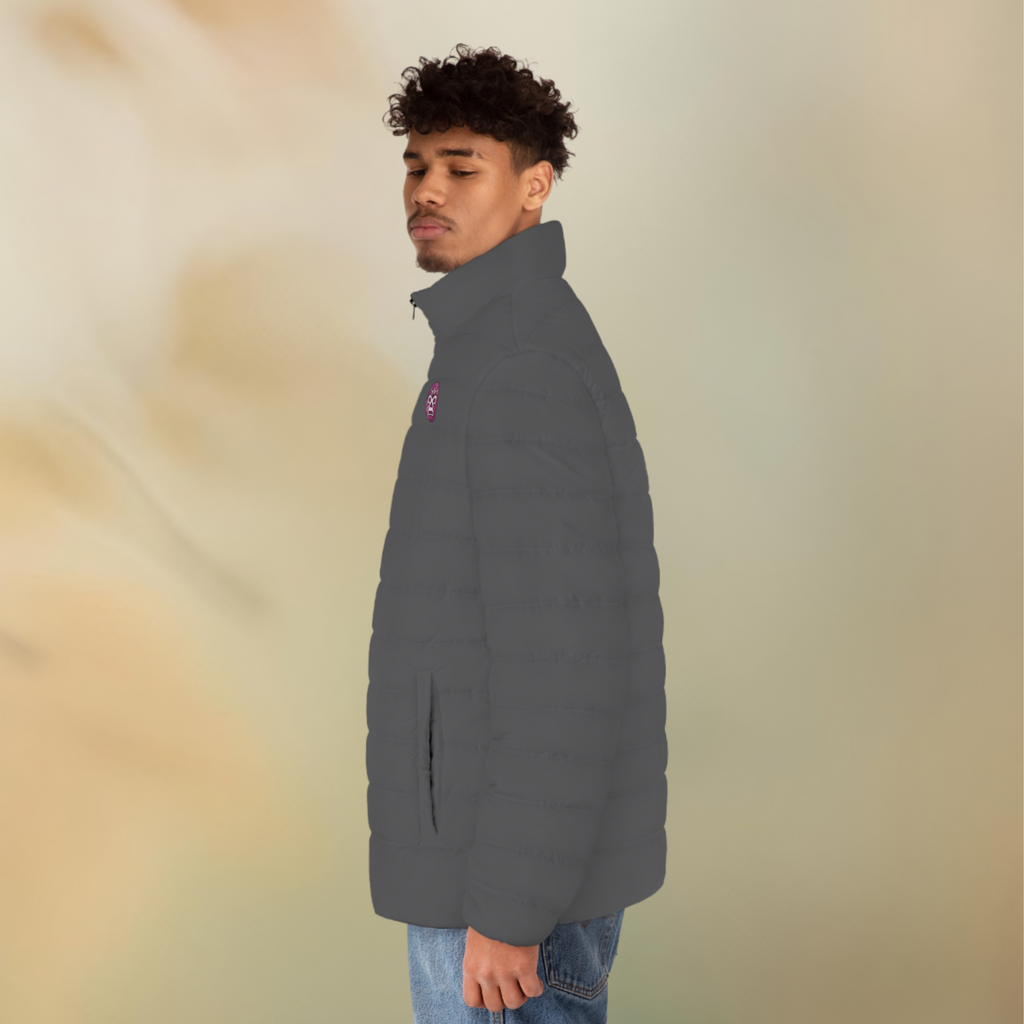Ein männliches Model zeigt sich von der linken Seite tragend die  Herren-Pufferjacke in einem Grey Stone Farbton. Die Jacke ist horizontal gesteppt, hat eine glatte Oberfläche, einen hohen Stehkragen und einen durchgehenden Reißverschluss. Auf der lingen Brusthöhe befindet sich ein Maori Emblem. Designed, by Just a Girl


