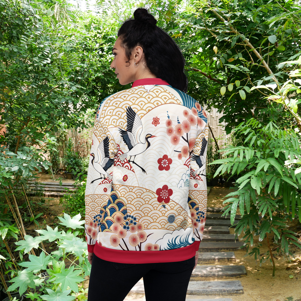Ein weibliches Model steht mit der Rückenansicht in einem Gartenumfeld und trägt eine kunstvoll designte Jacke mit japanischen inspiriertem Muster. Die Jacke zeigt florale Kirschblüten, stilisierte Wellen, goldene Fächer Elemente und einen Kranich auf Beigen Wellen Hintergrund.  By Just a Girl.
