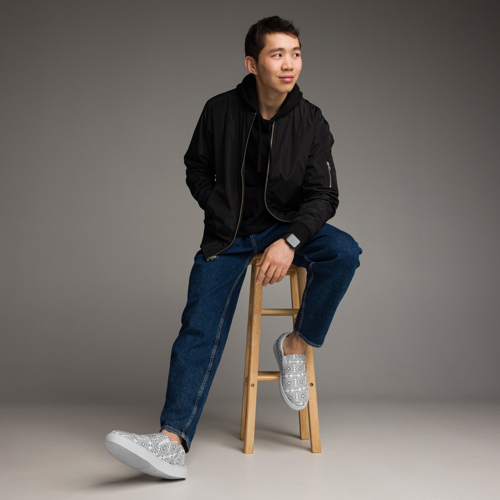 Ein Paar weißer Slip-On Canvas Schuhe mit einem wiederholten Muster aus unregelmäßigen, handgezeichnet wirkenden schwarzen Quadraten und Linien. Die Sohle ist weiß. Die Schuhe sind leicht nach links gedreht, sodass die Vorderseite beider Schuhe sichtbar ist.