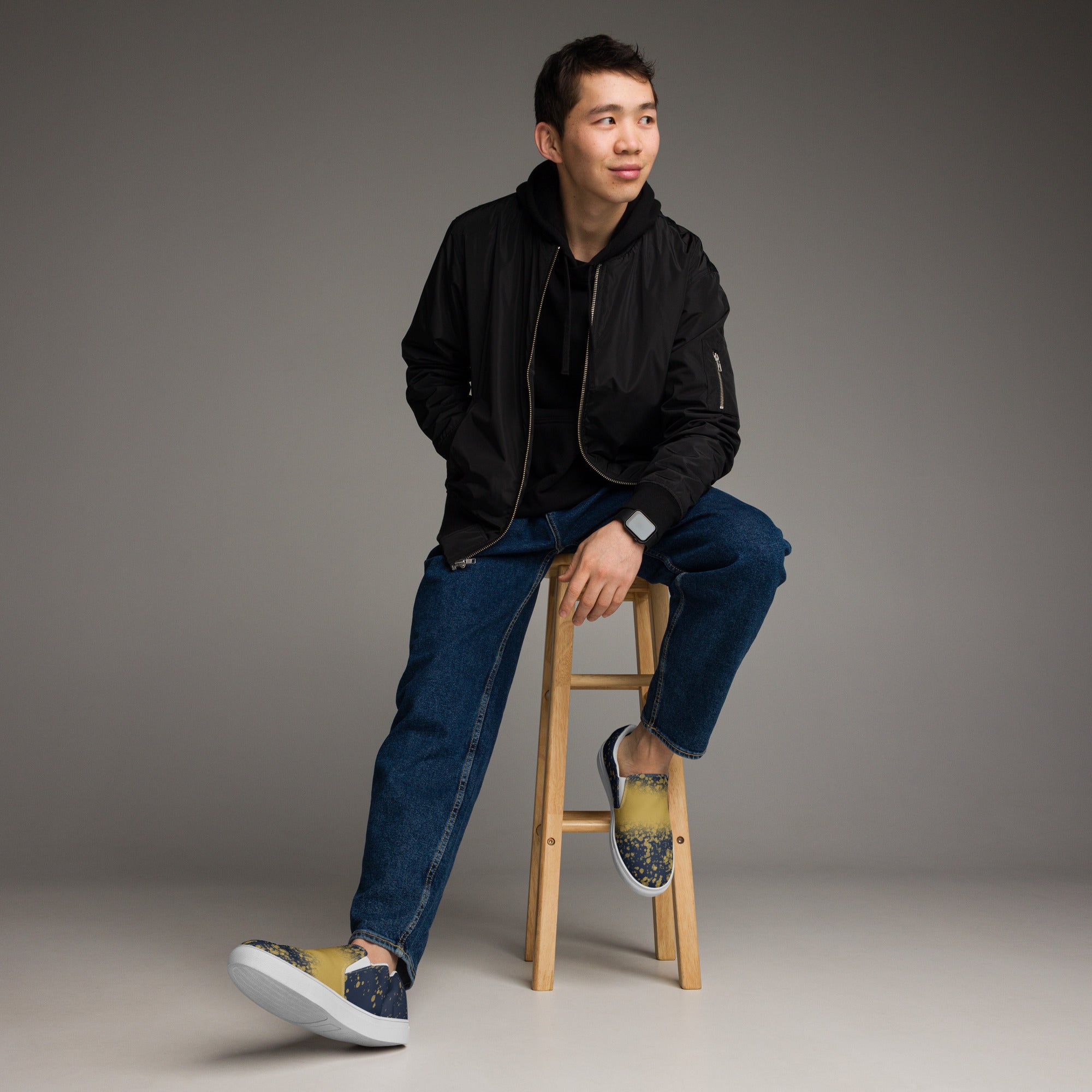 Ein Paar dunkelblauer Slip-On Canvas Schuhe mit einem Muster aus goldenen Farbspritzern, die von der Mitte des Schuhs nach außen hin weniger werden. Die Sohle ist weiß. Die Schuhe sind leicht nach links gedreht, sodass die Vorderseite beider Schuhe sichtbar ist.