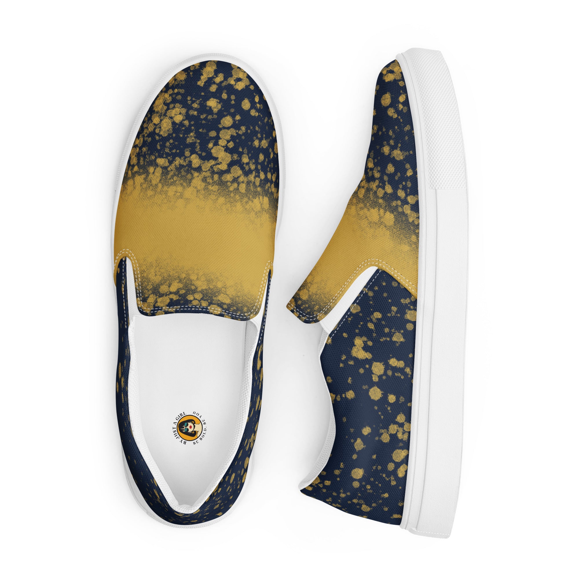 Ein Paar dunkelblauer Slip-On Canvas Schuhe mit einem Muster aus goldenen Farbspritzern, die von der Mitte des Schuhs nach außen hin weniger werden. Die Sohle ist weiß. Die Schuhe sind leicht nach links gedreht, sodass die Vorderseite beider Schuhe sichtbar ist.