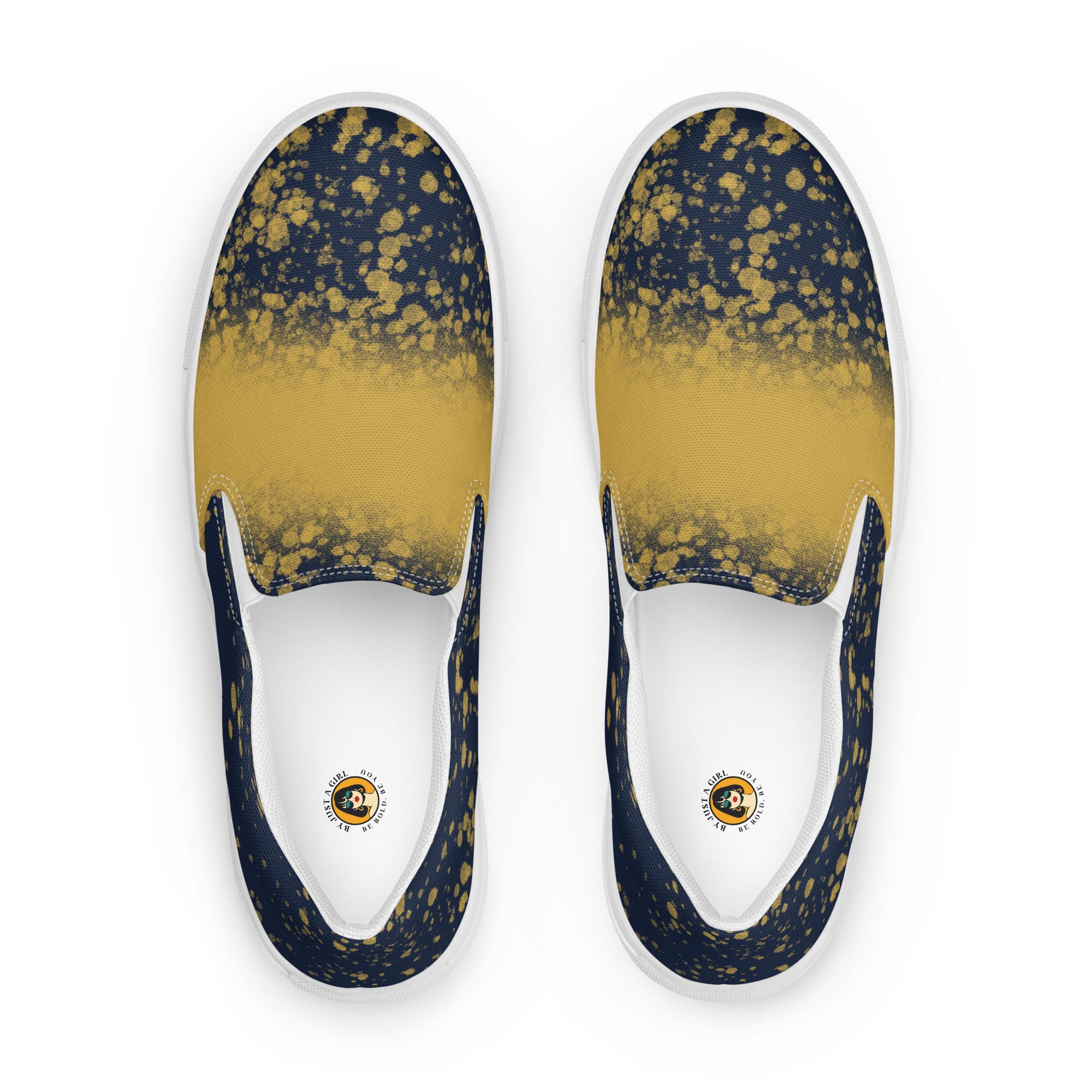 Ein Paar dunkelblauer Slip-On Canvas Schuhe mit einem Muster aus goldenen Farbspritzern, die von der Mitte des Schuhs nach außen hin weniger werden. Die Sohle ist weiß. Die Schuhe sind leicht nach links gedreht, sodass die Vorderseite beider Schuhe sichtbar ist.