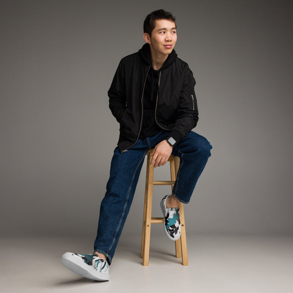 Ein Paar weißer Slip-On Canvas Schuhe mit einem abstrakten Muster aus großen, unregelmäßigen Klecksen und Flecken in Schwarz, Grau und leuchtendem Türkis. Die Sohle ist weiß. Die Schuhe sind leicht nach links gedreht, sodass die Vorderseite beider Schuhe sichtbar ist.