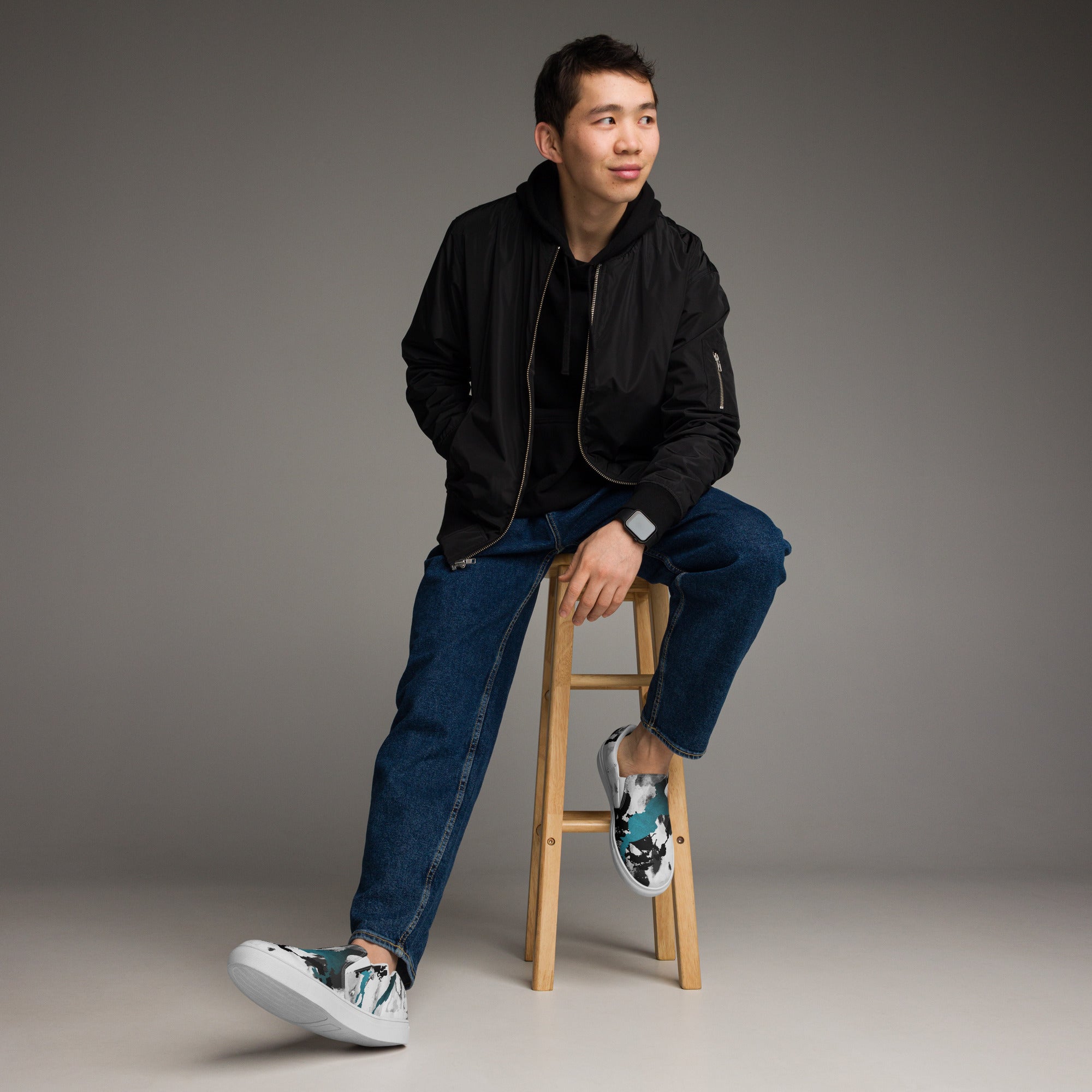 Ein Paar weißer Slip-On Canvas Schuhe mit einem abstrakten Muster aus großen, unregelmäßigen Klecksen und Flecken in Schwarz, Grau und leuchtendem Türkis. Die Sohle ist weiß. Die Schuhe sind leicht nach links gedreht, sodass die Vorderseite beider Schuhe sichtbar ist.