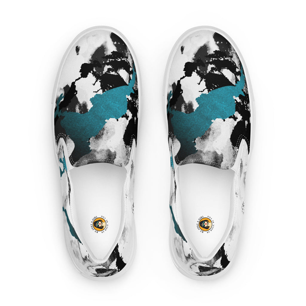 Ein Paar weißer Slip-On Canvas Schuhe mit einem abstrakten Muster aus großen, unregelmäßigen Klecksen und Flecken in Schwarz, Grau und leuchtendem Türkis. Die Sohle ist weiß. Die Schuhe sind leicht nach links gedreht, sodass die Vorderseite beider Schuhe sichtbar ist.