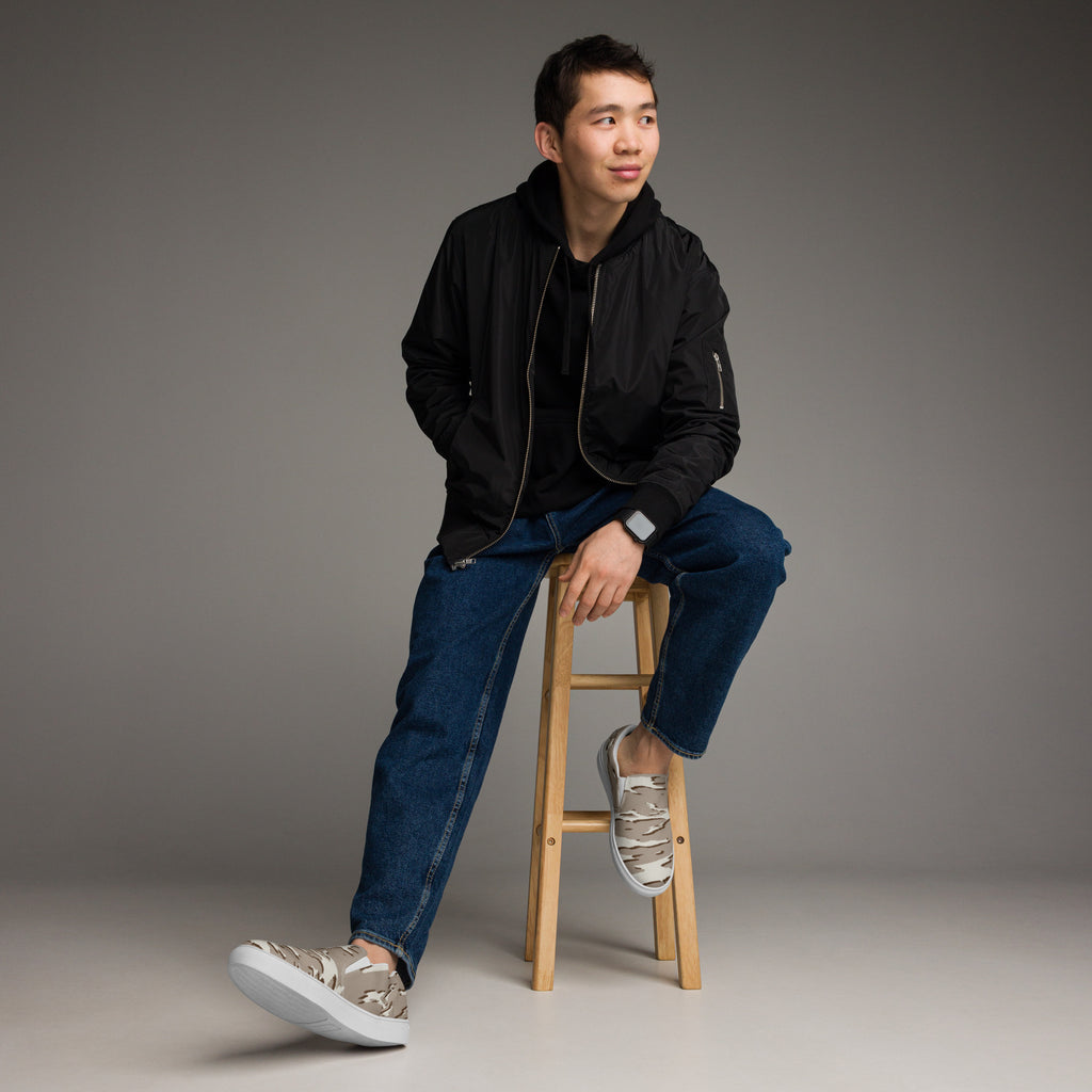 Ein Paar beiger Slip-On Canvas Schuhe mit einem wellenförmigen Camouflage-Muster in Hellbeige und Dunkelbeige. Die Sohle ist weiß. Die Schuhe sind leicht nach links gedreht, sodass die Vorderseite beider Schuhe sichtbar ist.