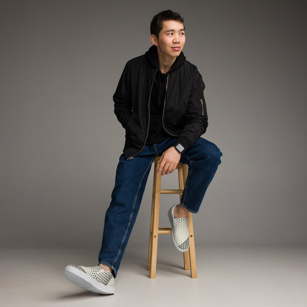 Ein Paar cremefarbener Slip-On Canvas Schuhe mit einem Muster aus schwarzen Punkten. Die Punkte sind am Rand des Schuhs größer und werden zur Mitte hin kleiner. Die Sohle ist weiß. Die Schuhe sind leicht nach links gedreht, sodass die Vorderseite beider Schuhe sichtbar ist.