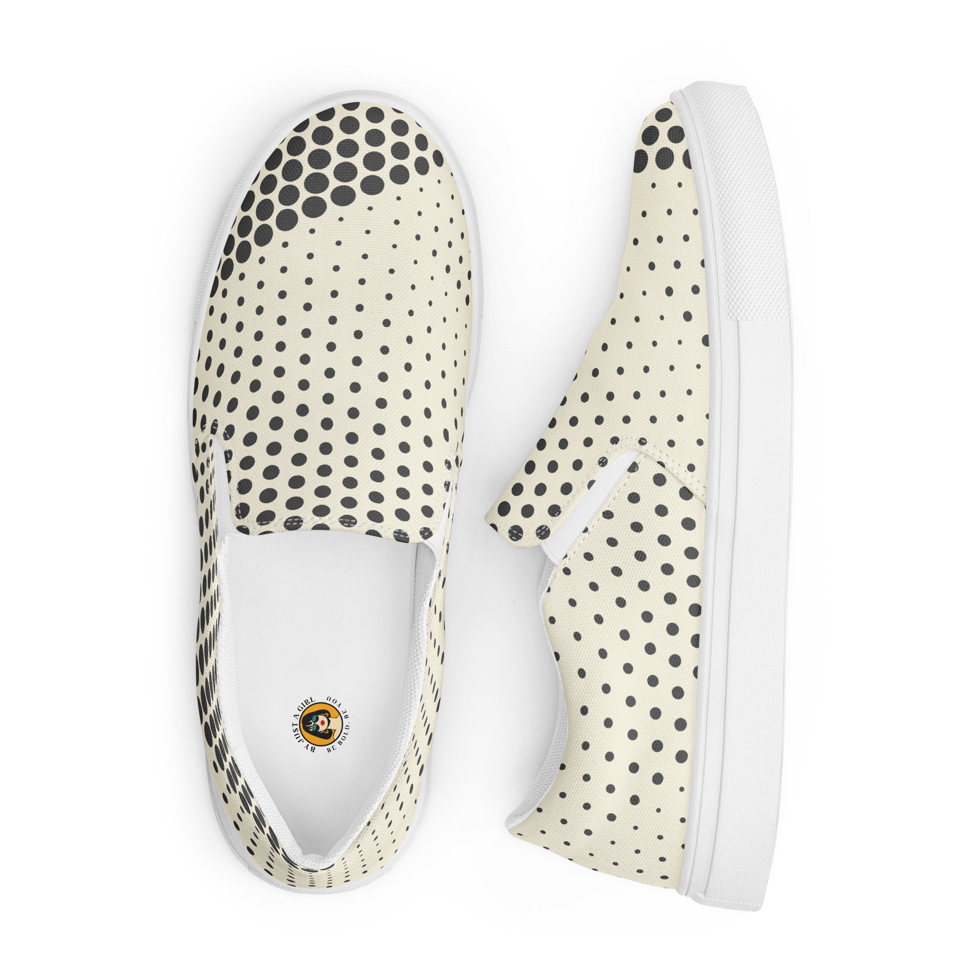 Ein Paar cremefarbener Slip-On Canvas Schuhe mit einem Muster aus schwarzen Punkten. Die Punkte sind am Rand des Schuhs größer und werden zur Mitte hin kleiner. Die Sohle ist weiß. Die Schuhe sind leicht nach links gedreht, sodass die Vorderseite beider Schuhe sichtbar ist.