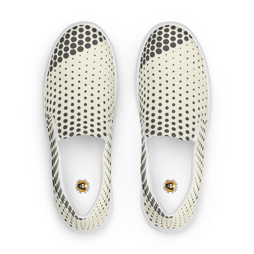 Ein Paar cremefarbener Slip-On Canvas Schuhe mit einem Muster aus schwarzen Punkten. Die Punkte sind am Rand des Schuhs größer und werden zur Mitte hin kleiner. Die Sohle ist weiß. Die Schuhe sind leicht nach links gedreht, sodass die Vorderseite beider Schuhe sichtbar ist.