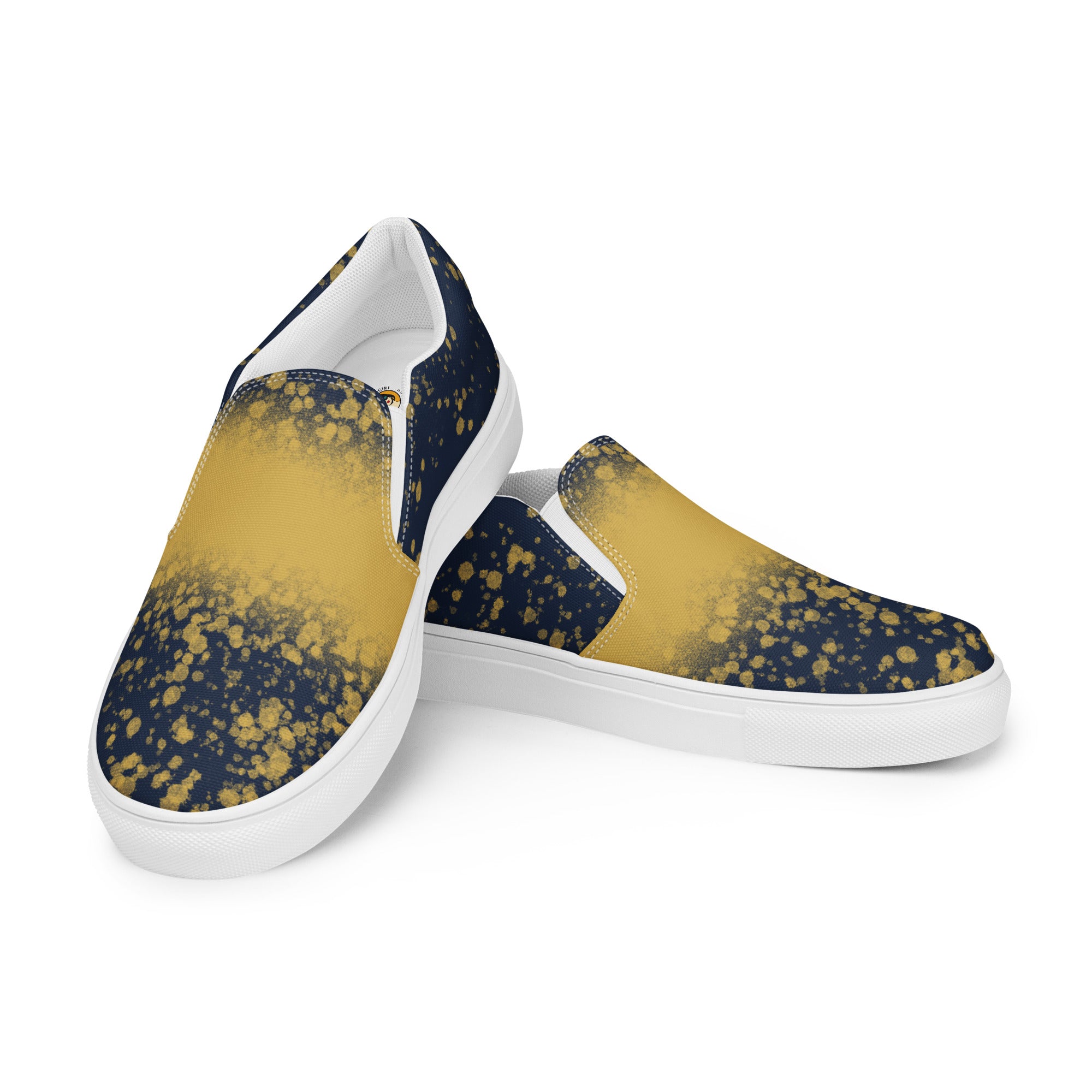 Ein Paar dunkelblauer Slip-On Canvas Schuhe mit einem Muster aus goldenen Farbspritzern, die von der Mitte des Schuhs nach außen hin weniger werden. Die Sohle ist weiß. Die Schuhe sind leicht nach links gedreht, sodass die Vorderseite beider Schuhe sichtbar ist.