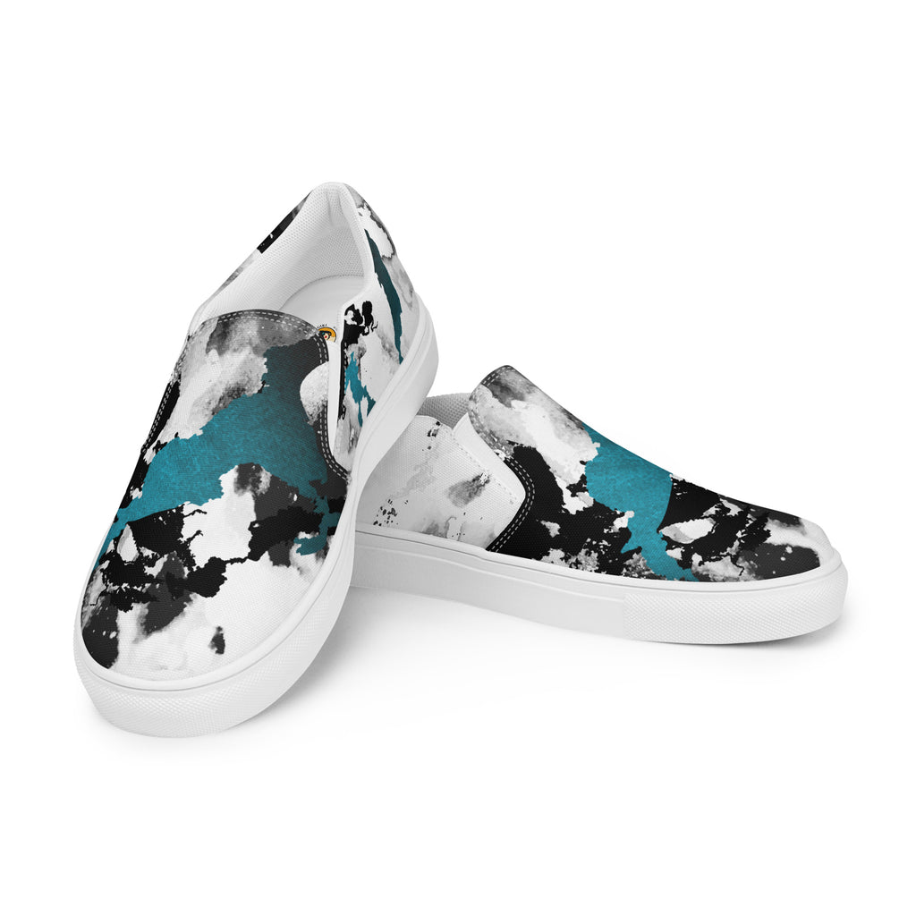 Ein Paar weißer Slip-On Canvas Schuhe mit einem abstrakten Muster aus großen, unregelmäßigen Klecksen und Flecken in Schwarz, Grau und leuchtendem Türkis. Die Sohle ist weiß. Die Schuhe sind leicht nach links gedreht, sodass die Vorderseite beider Schuhe sichtbar ist.