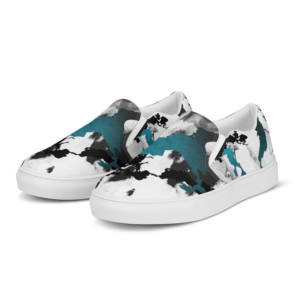 Ein Paar weißer Slip-On Canvas Schuhe mit einem abstrakten Muster aus großen, unregelmäßigen Klecksen und Flecken in Schwarz, Grau und leuchtendem Türkis. Die Sohle ist weiß. Die Schuhe sind leicht nach links gedreht, sodass die Vorderseite beider Schuhe sichtbar ist.