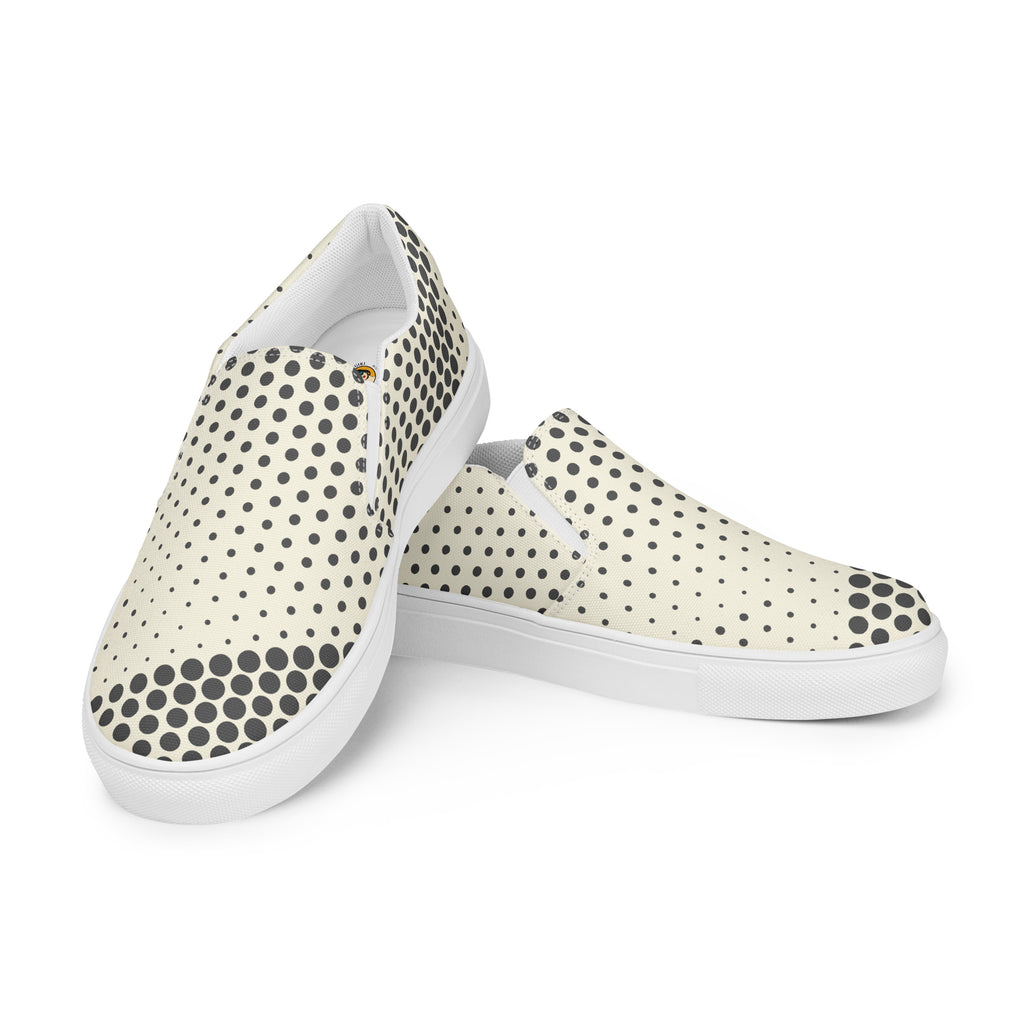 Ein Paar cremefarbener Slip-On Canvas Schuhe mit einem Muster aus schwarzen Punkten. Die Punkte sind am Rand des Schuhs größer und werden zur Mitte hin kleiner. Die Sohle ist weiß. Die Schuhe sind leicht nach links gedreht, sodass die Vorderseite beider Schuhe sichtbar ist.