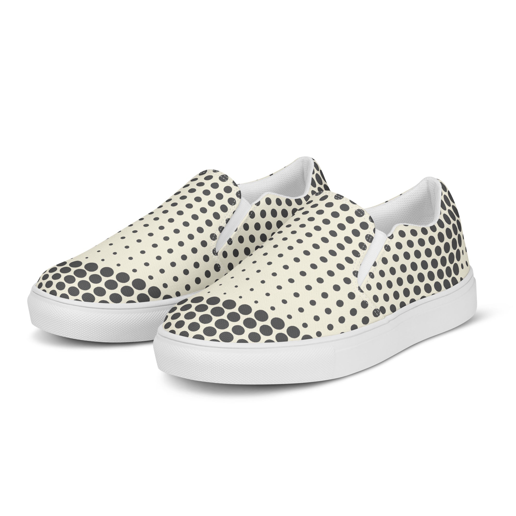Ein Paar cremefarbener Slip-On Canvas Schuhe mit einem Muster aus schwarzen Punkten. Die Punkte sind am Rand des Schuhs größer und werden zur Mitte hin kleiner. Die Sohle ist weiß. Die Schuhe sind leicht nach links gedreht, sodass die Vorderseite beider Schuhe sichtbar ist.