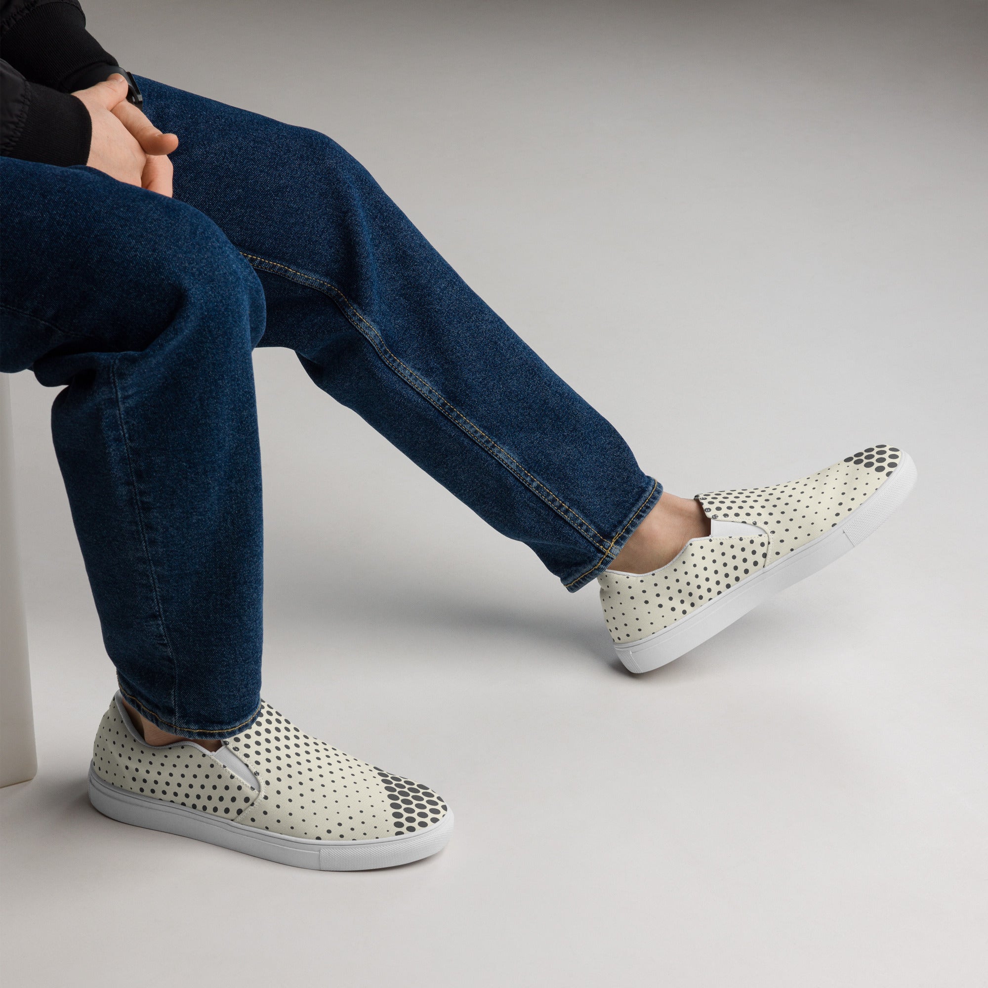 Ein Paar cremefarbener Slip-On Canvas Schuhe mit einem Muster aus schwarzen Punkten. Die Punkte sind am Rand des Schuhs größer und werden zur Mitte hin kleiner. Die Sohle ist weiß. Die Schuhe sind leicht nach links gedreht, sodass die Vorderseite beider Schuhe sichtbar ist.