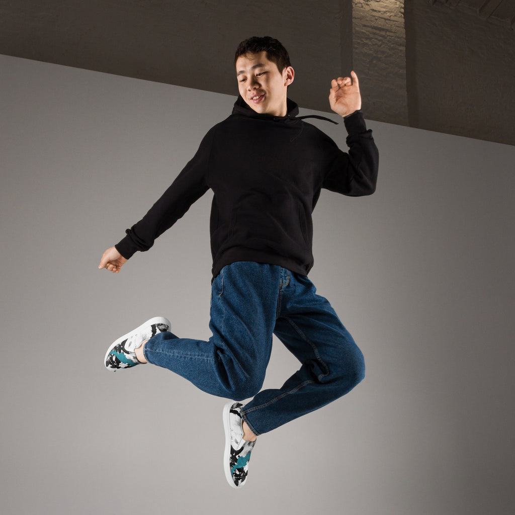 Ein Paar weißer Slip-On Canvas Schuhe mit einem abstrakten Muster aus großen, unregelmäßigen Klecksen und Flecken in Schwarz, Grau und leuchtendem Türkis. Die Sohle ist weiß. Die Schuhe sind leicht nach links gedreht, sodass die Vorderseite beider Schuhe sichtbar ist.