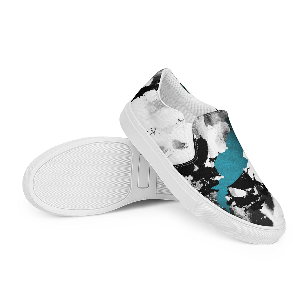 Ein Paar weißer Slip-On Canvas Schuhe mit einem abstrakten Muster aus großen, unregelmäßigen Klecksen und Flecken in Schwarz, Grau und leuchtendem Türkis. Die Sohle ist weiß. Die Schuhe sind leicht nach links gedreht, sodass die Vorderseite beider Schuhe sichtbar ist.