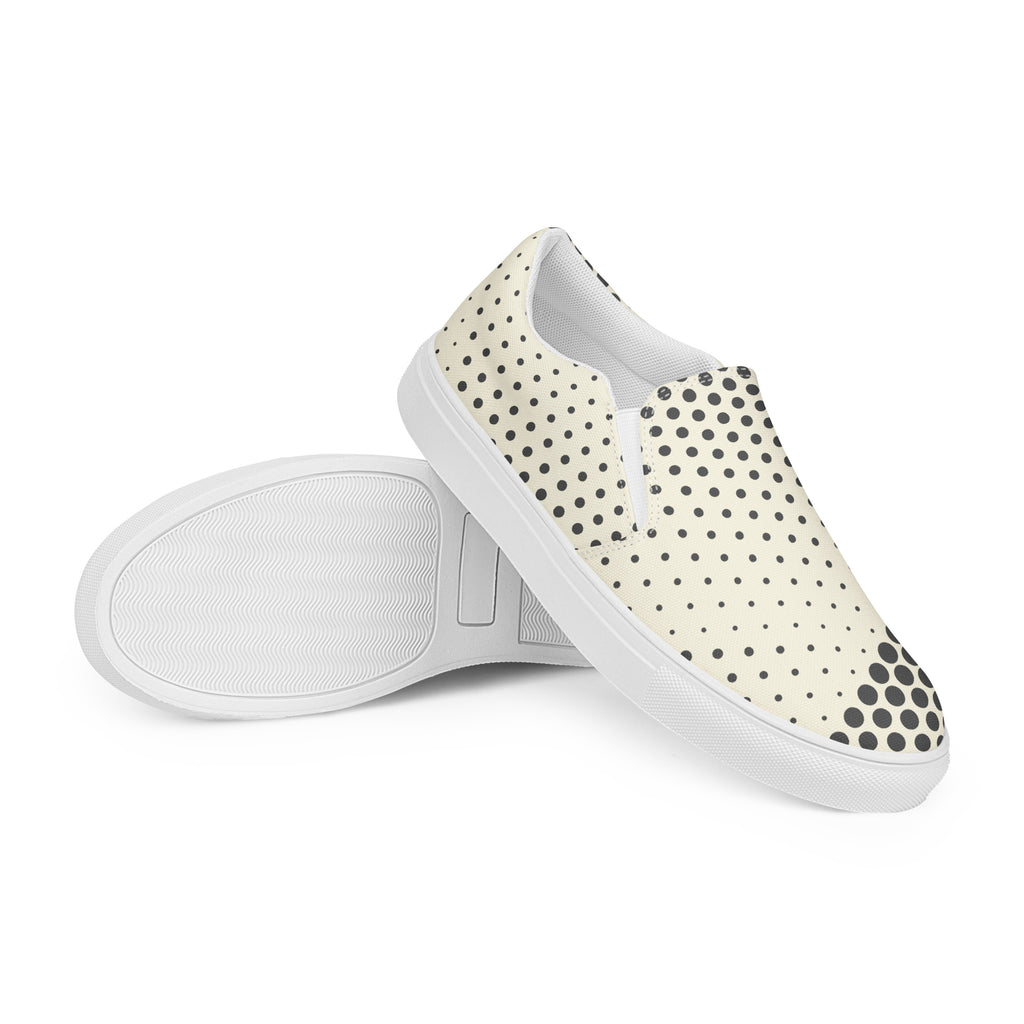 Ein Paar cremefarbener Slip-On Canvas Schuhe mit einem Muster aus schwarzen Punkten. Die Punkte sind am Rand des Schuhs größer und werden zur Mitte hin kleiner. Die Sohle ist weiß. Die Schuhe sind leicht nach links gedreht, sodass die Vorderseite beider Schuhe sichtbar ist.