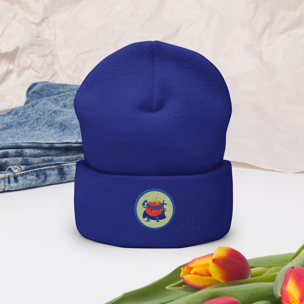 Ein Royal Blaues Cuffed Beanie mit aufgesticktem Rotem Tomaten Superhelden Gesicht (Cartoon) in einem runden blauen Rahmen auf der Vorderseite, präsentiert zur Dekoration mit Jeans und Blumen. Designed, by Just a Girl.
