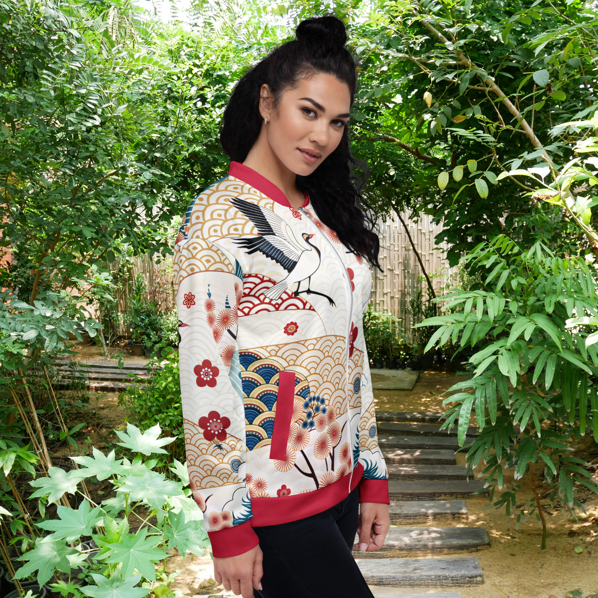 Das weibliches Model steht mit seitlicher Ansicht in einem Gartenumfeld und trägt eine kunstvoll designte Jacke mit japanischen inspiriertem Muster. Die Jacke zeigt florale Kirschblüten, stilisierte Wellen, goldene Fächer Elemente und einen Kranich auf Beigen Wellen Hintergrund mit roten Akzenten an Kragen, Bündchen und Taschen. By Just a Girl.