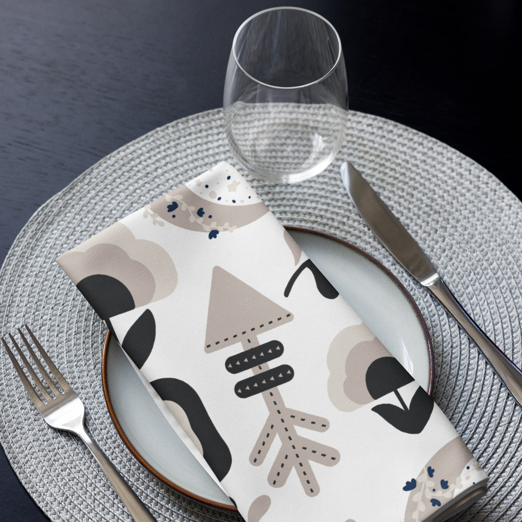 Eine Stoff Serviette gefaltete auf einem gedecktem Teller. Das Muster zeigt ein abstraktes Design mit Boho-Elementen wie stilisierten Pfeilen, einem Regenbogen, einer gepunkteten Mondsichel und floralen Motiven in Schwarz, Beige und Brauntönen.