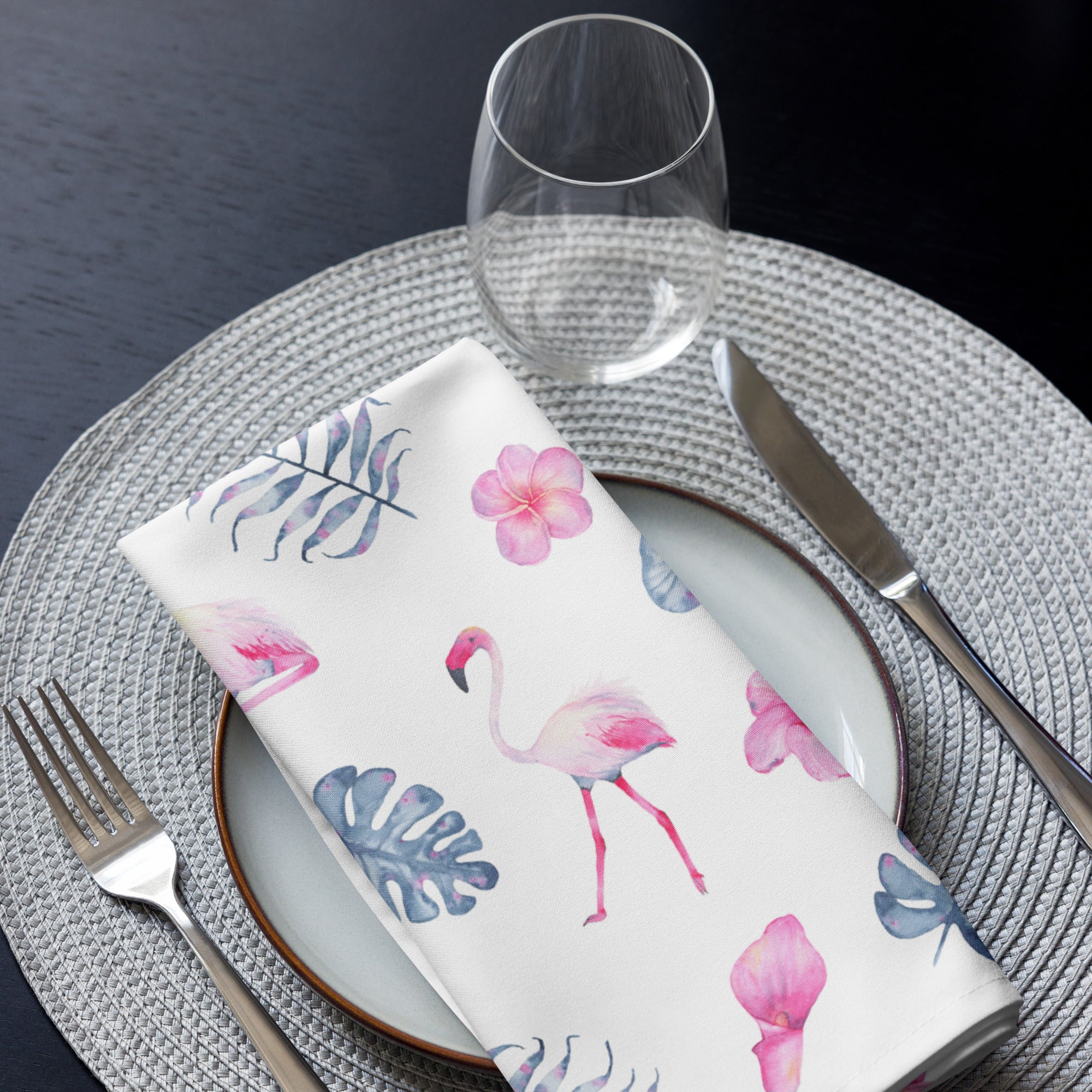 Eine Stoff Serviette gefaltete auf einem gedecktem Teller. Der Hintergrund der Servietten ist weiß und zeigt ein tropisches Muster aus rosa Flamingos, pinkfarbenen Hibiskusblüten und blaugrauen, großen Blättern in einem Aquarell-Stil.