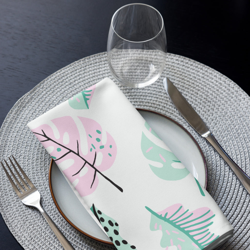 Eine Stoff Serviette gefaltete auf einem gedecktem Teller. Der Hintergrund der Servietten ist weiß und zeigt ein modernes, abstraktes Muster aus Blättern in zarten Rosa- und Mintgrün-Tönen, ergänzt durch kleine schwarze Akzente.
