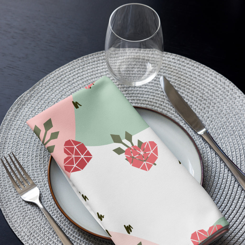 Eine Stoff Serviette gefaltete auf einem gedecktem Teller. Der Hintergrund ist weiß und zeigt ein abstraktes Muster mit rosafarbenen Herzen, hellgrünen Blattformen und kleinen schwarzen Strichen.
