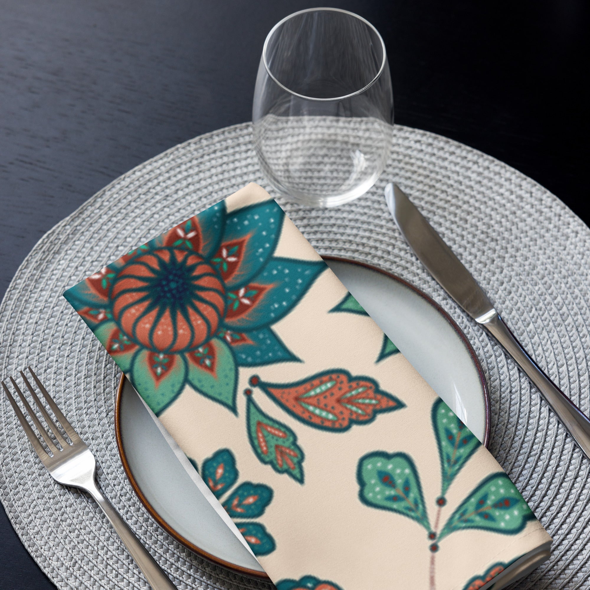 Eine Stoff Serviette gefaltete auf einem gedecktem Teller. Der Hintergrund der Servietten ist Beige und zeigt ein ornamentales Muster aus stilisierten Vögeln, die auf floralen Zweigen mit Blumen und Blättern sitzen. Die Motive sind in Türkis-, Rostrot- und Brauntönen gehalten.