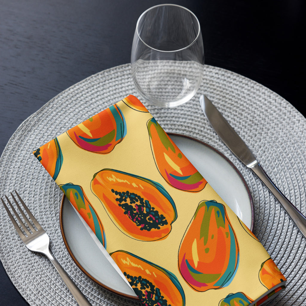 Eine Stoff Serviette gefaltete auf einem gedecktem Teller. Das Muster zeigt eine Mischung aus ganzen und halben Papayas in leuchtenden Orange-, Rot- und Blautönen mit schwarzen Kernen. Der Hintergrund der Servietten ist einfarbig gelb.