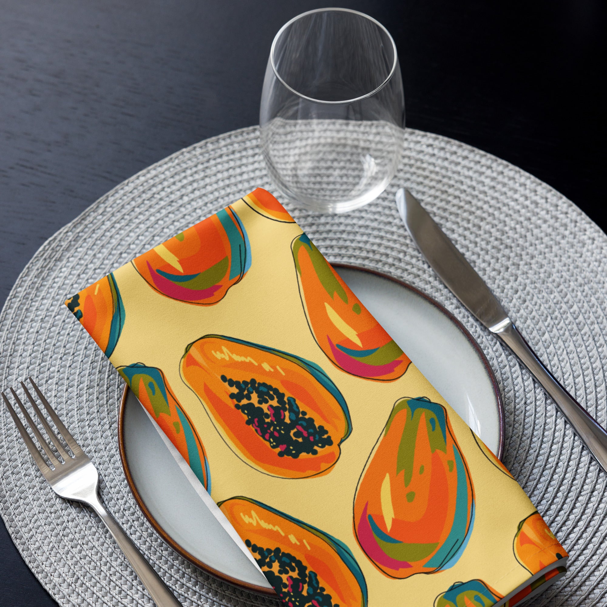 Eine Stoff Serviette gefaltete auf einem gedecktem Teller. Das Muster zeigt eine Mischung aus ganzen und halben Papayas in leuchtenden Orange-, Rot- und Blautönen mit schwarzen Kernen. Der Hintergrund der Servietten ist einfarbig gelb.