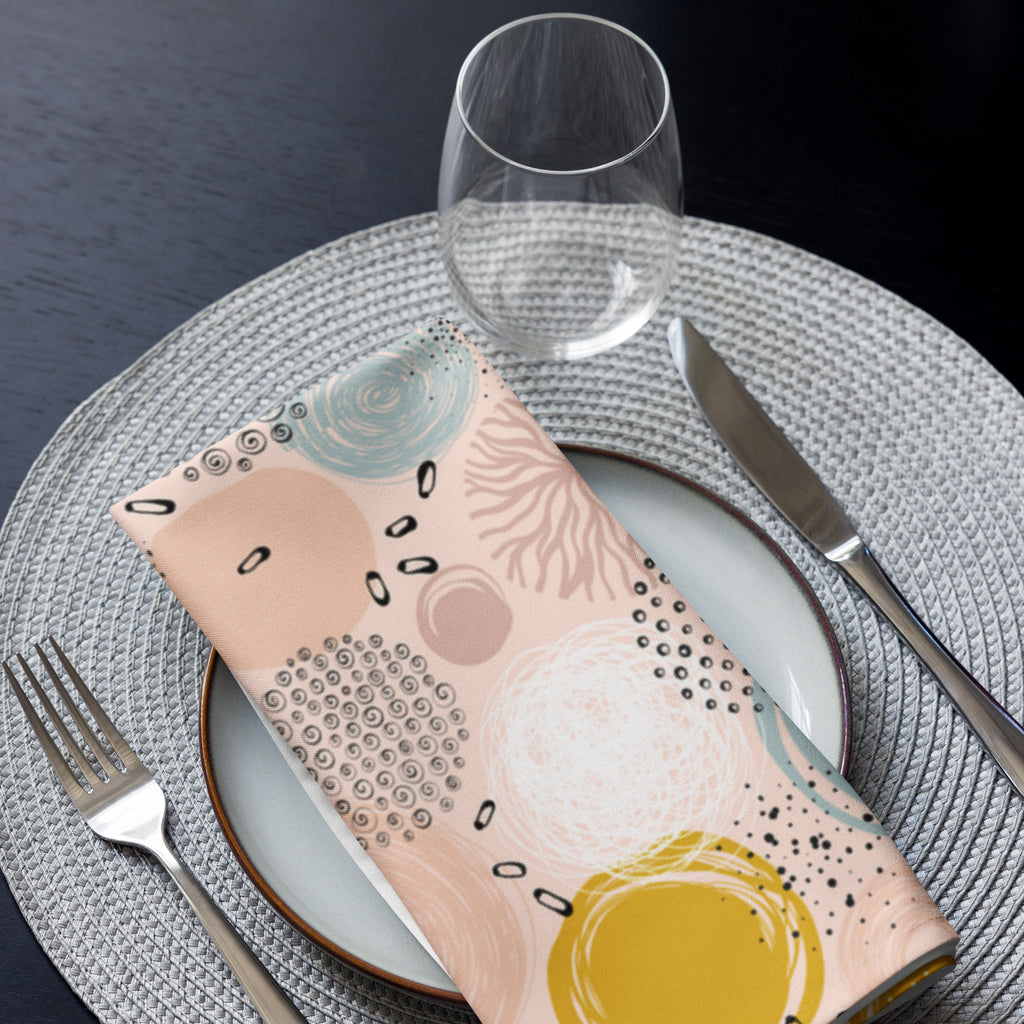 Eine Stoff Serviette gefaltete auf einem gedecktem Teller. Die Servietten haben einen rosafarbenen Hintergrund und zeigen ein abstraktes Muster aus Kreisen in Gelb, Blau und Weiß sowie kleine schwarze Sprenkel.