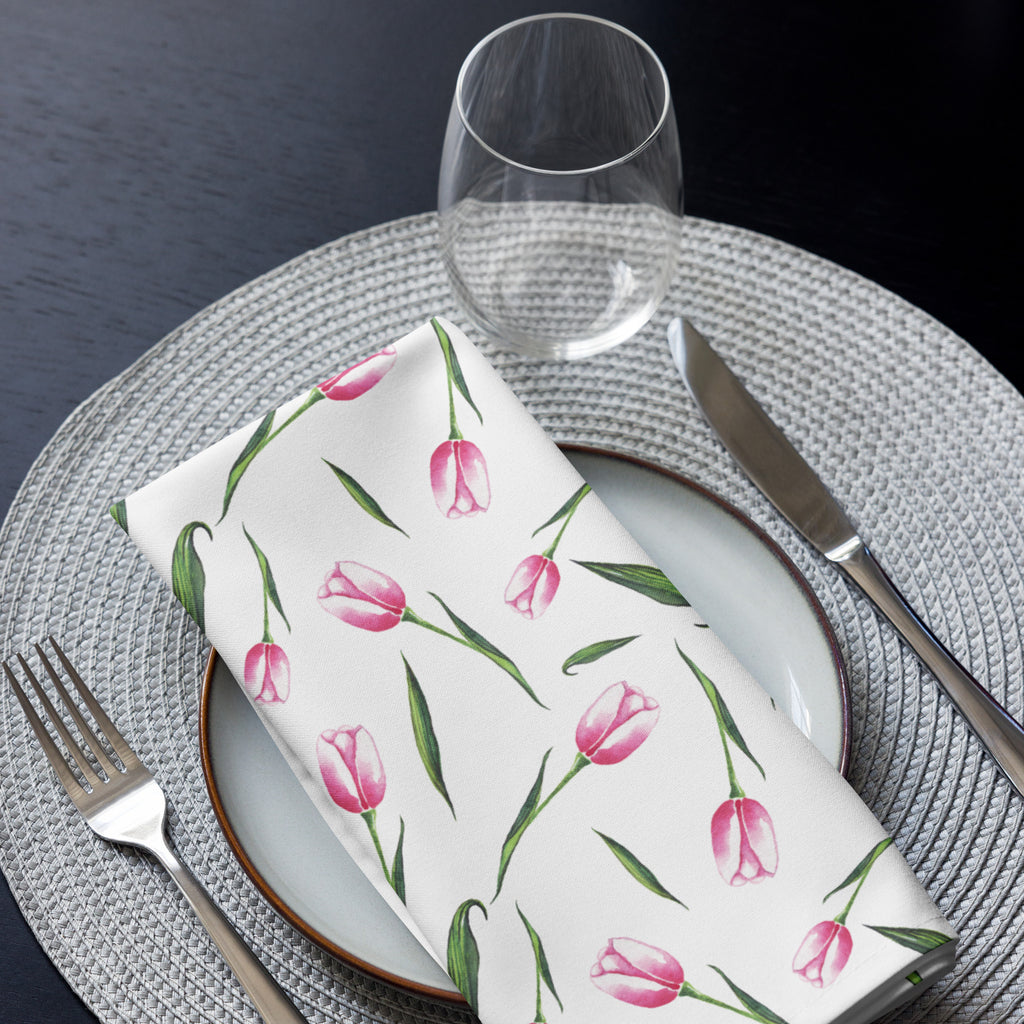 Eine Stoff Serviette gefaltete auf einem gedecktem Teller. Der weiße Hintergrund ist mit einer Streuung aus zarten, rosa Tulpen bedruckt, deren Stängel und Blätter in frischem Grün gehalten sind.