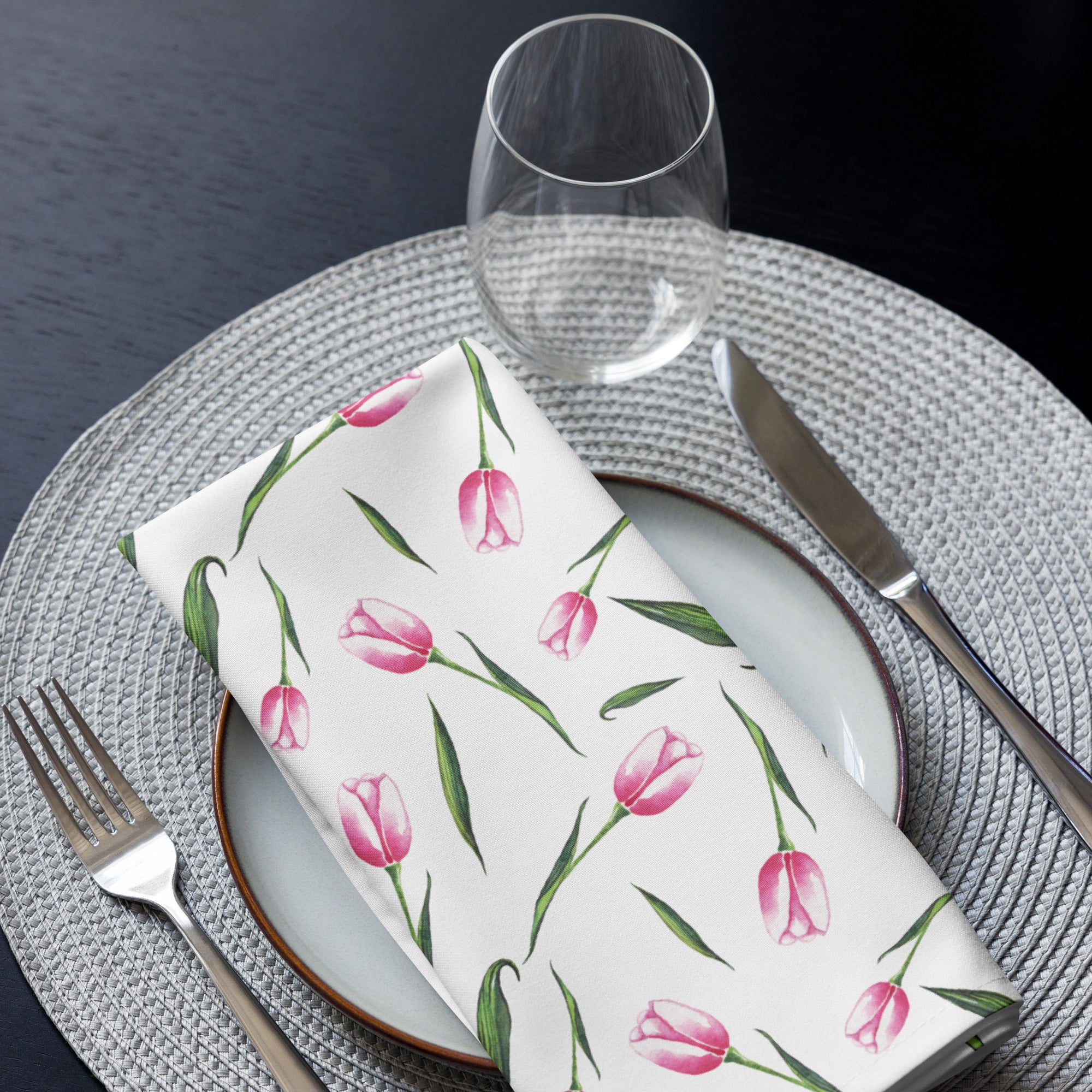 Eine Stoff Serviette gefaltete auf einem gedecktem Teller. Der weiße Hintergrund ist mit einer Streuung aus zarten, rosa Tulpen bedruckt, deren Stängel und Blätter in frischem Grün gehalten sind.