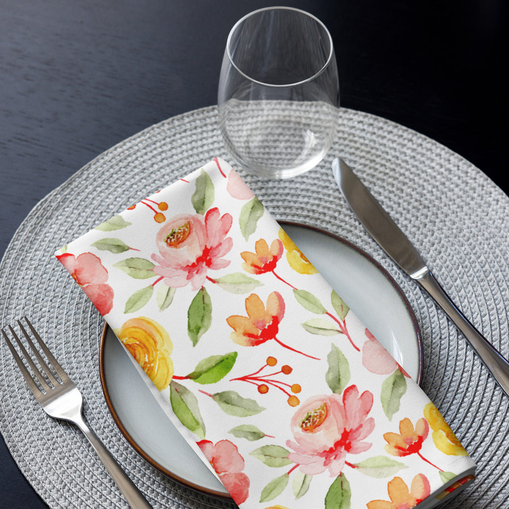 Eine Stoff Serviette gefaltete auf einem gedecktem Teller. Der Hintergrund der Servietten ist weiß und zeigt ein florales Muster aus gelben Rosen mit grünen Blättern sowie rosafarbene Blumen mit leicht roten Akzenten.