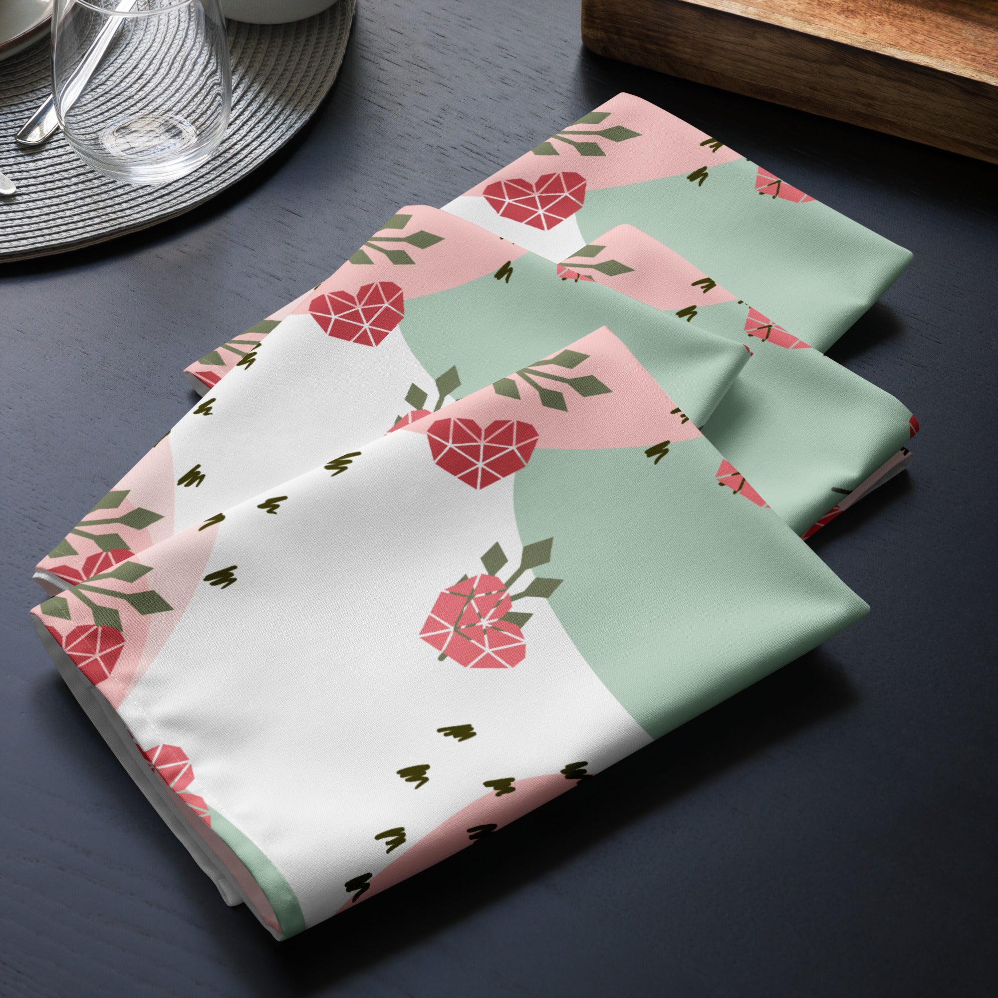 4er Set Stoff Servietten gefaltet auf dem Tisch platziert. Der Hintergrund ist weiß und zeigt ein abstraktes Muster mit rosafarbenen Herzen, hellgrünen Blattformen und kleinen schwarzen Strichen.