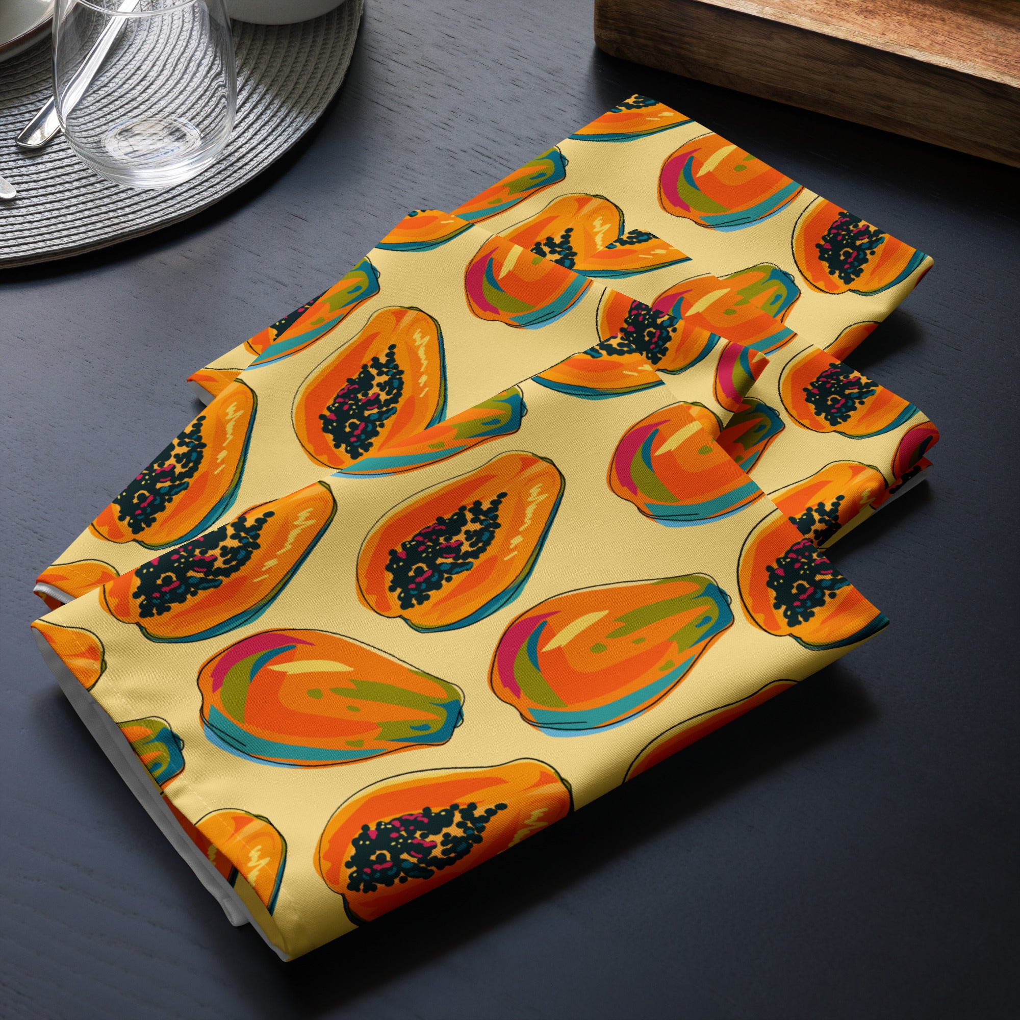 4er Set Stoff Servietten gefaltet auf dem Tisch platziert. Das Muster zeigt eine Mischung aus ganzen und halben Papayas in leuchtenden Orange-, Rot- und Blautönen mit schwarzen Kernen. Der Hintergrund der Servietten ist einfarbig gelb.