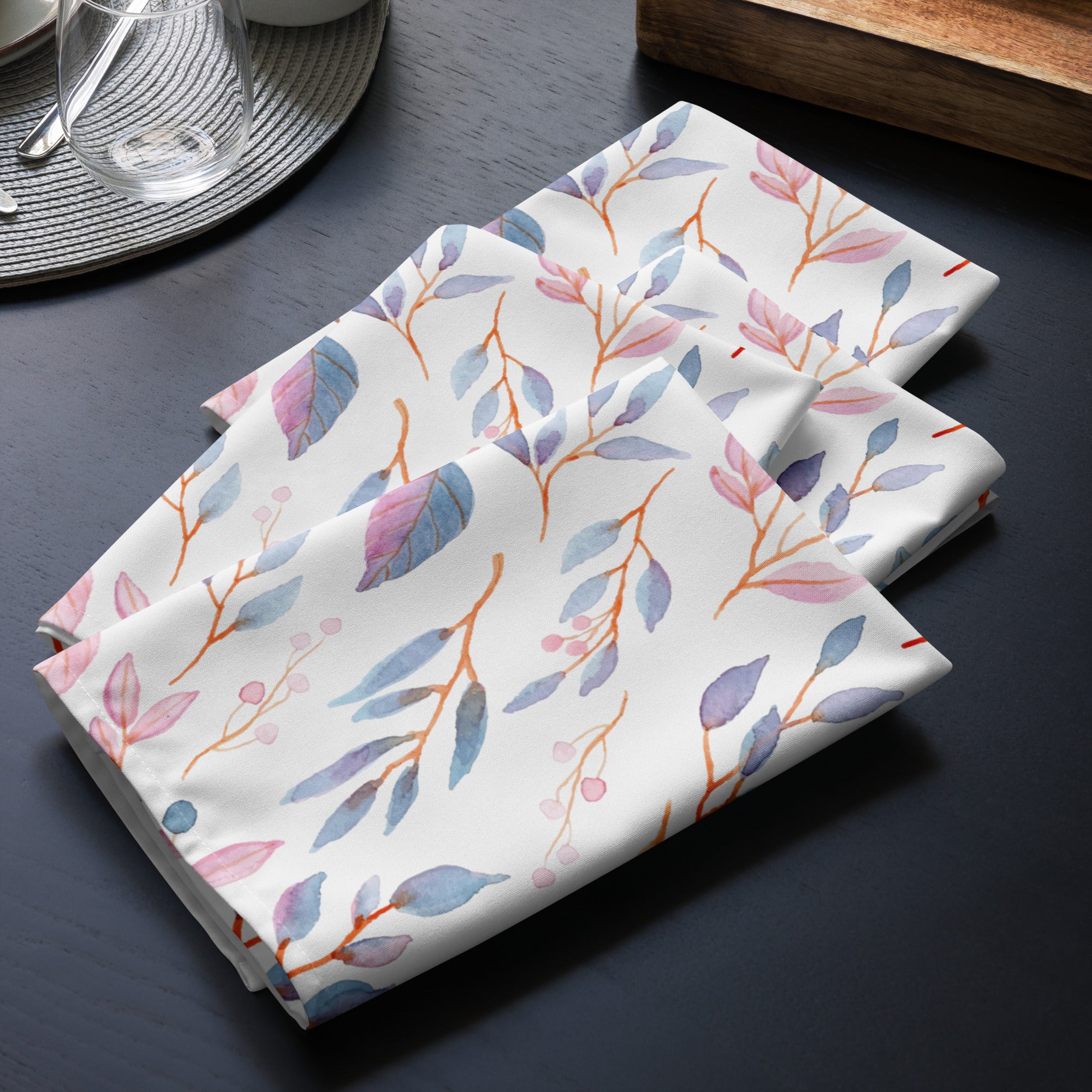 4er Set Stoff Servietten gefaltet auf dem Tisch platziert. Der Hintergrund der Servietten ist weiß und zeigt ein filigranes Muster aus Zweigen mit Blättern und kleinen Beeren. Die Blätter sind in zarten Blau-, Lila- und Rosatönen gehalten, während die Beeren dunkelblau sind.