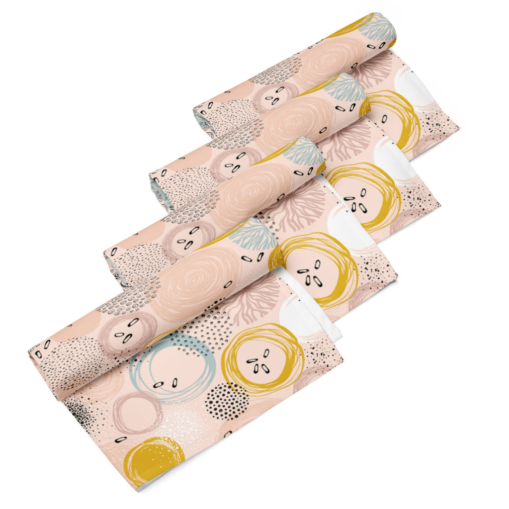 Ein Set aus vier gerollten Stoff-Servietten, freigestellt auf weißem Hintergrund. Die Servietten haben einen rosafarbenen Hintergrund und zeigen ein abstraktes Muster aus Kreisen in Gelb, Blau und Weiß sowie kleine schwarze Sprenkel.