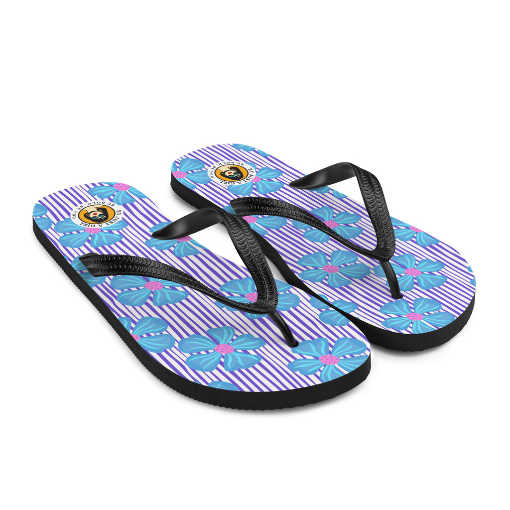 Ein Paar Flip-Flops mit einem Obermaterial, das ein Muster aus vertikalen lila und weißen Streifen zeigt, überlagert von großen, stilisierten blauen Blüten mit pinkfarbenen Zentren. Die Riemen sind schwarz und haben eine strukturierte Oberfläche. Auf der Ferse jedes Flip-Flops befindet sich ein rundes gelbes Emblem mit dem Text "BY JUST A GIRL" und "BE BOLD. BE YOU" sowie dem stilisierten Kopf einer Frau mit Bob-Frisur und Sonnenbrille.