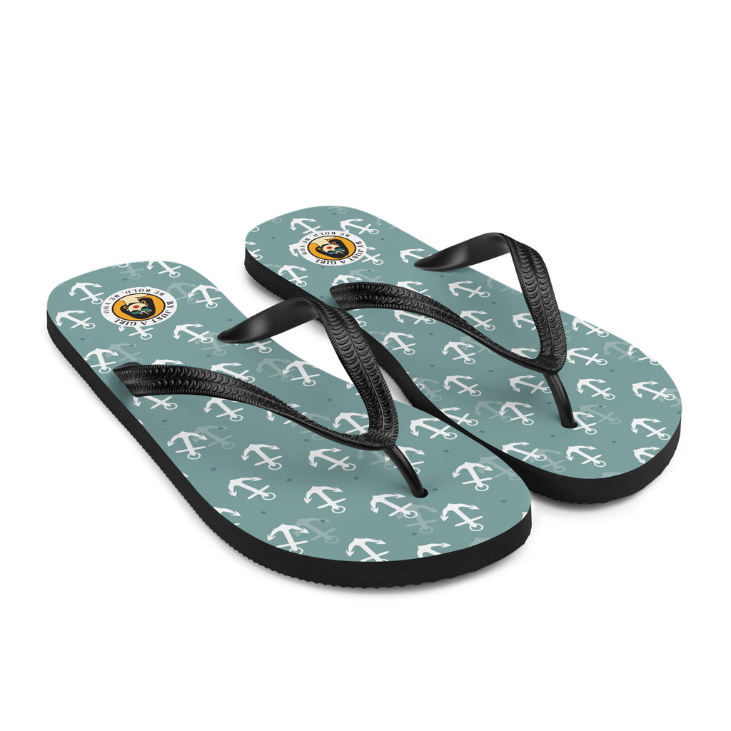 Ein Paar Flip-Flops mit einem Obermaterial in einem gedämpften Türkis- oder Blaugrünton, das mit einem wiederkehrenden Muster aus stilisierten weißen Ankern und kleinen grauen Punkten bedeckt ist. Die Riemen sind schwarz und haben eine strukturierte Oberfläche. Auf der Ferse jedes Flip-Flops befindet sich ein rundes gelbes Emblem mit dem Text "BY JUST A GIRL" und "BE BOLD. BE YOU" sowie dem stilisierten Kopf einer Frau mit Bob-Frisur und Sonnenbrille.