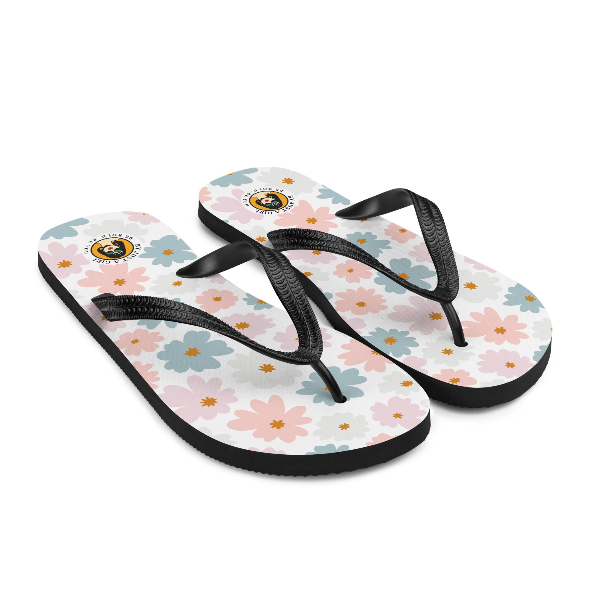 Ein Paar Flip-Flops mit einem weißen Obermaterial, das mit einem wiederkehrenden Muster aus stilisierten, fünfblättrigen Blüten in Pastellfarben wie Hellrosa, Hellblau und Beige bedeckt ist, ergänzt durch kleine orangefarbene Sterne in der Mitte einiger Blüten. Die Riemen sind schwarz und haben eine strukturierte Oberfläche. Auf der Ferse jedes Flip-Flops befindet sich ein rundes gelbes Emblem mit dem Text "BY JUST A GIRL" und "BE BOLD. BE YOU" sowie dem stilisierten Kopf einer Frau mit Bob-Frisur und Sonne