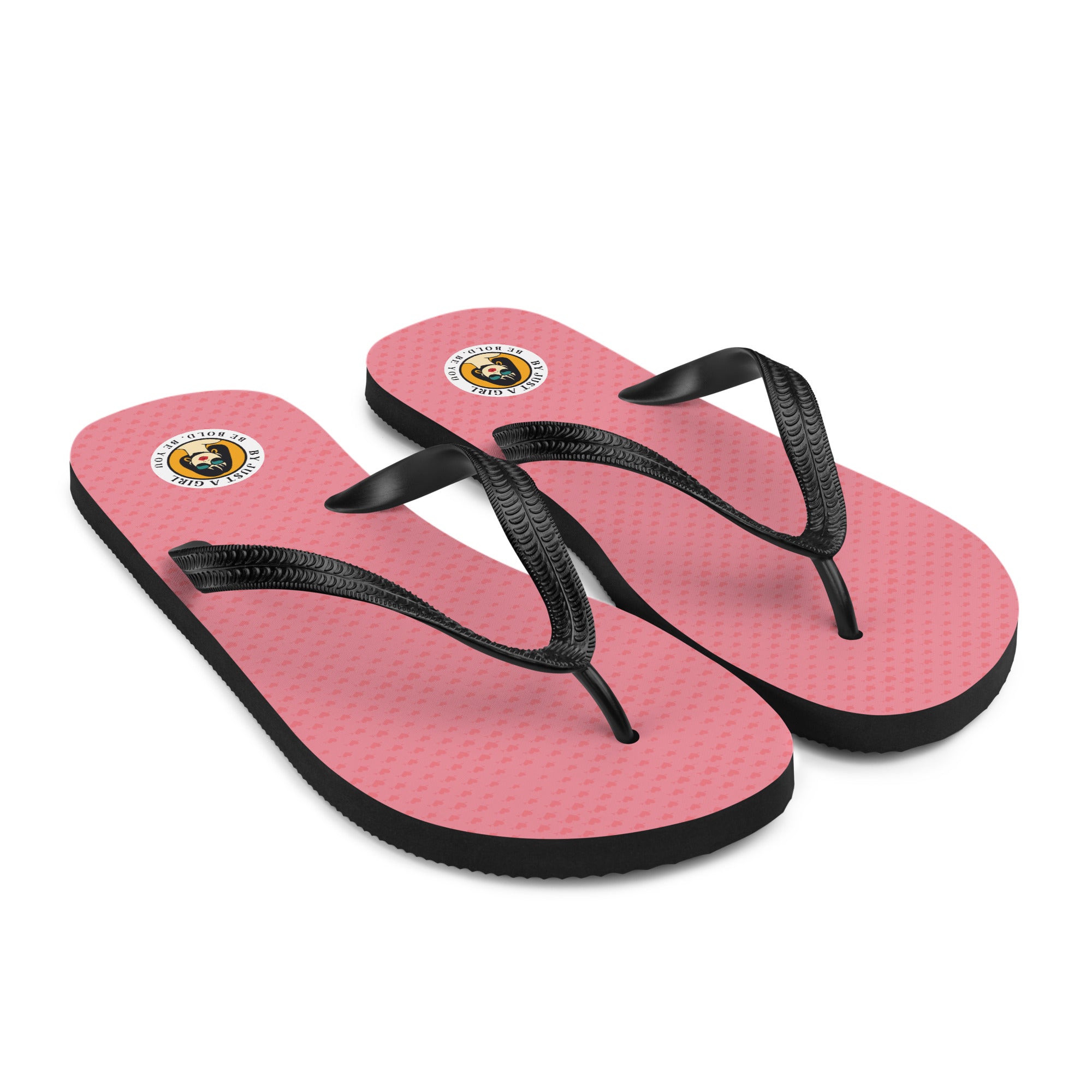 Ein Paar Flip-Flops mit einem Obermaterial, kleine Herzen in Rosa und Rot zeigt. Die Riemen sind schwarz und haben eine strukturierte Oberfläche. Auf der Ferse jedes Flip-Flops befindet sich ein rundes gelbes Emblem mit dem Text "BY JUST A GIRL" und "BE BOLD. BE YOU" sowie dem stilisierten Kopf einer Frau mit Bob-Frisur und Sonnenbrille.