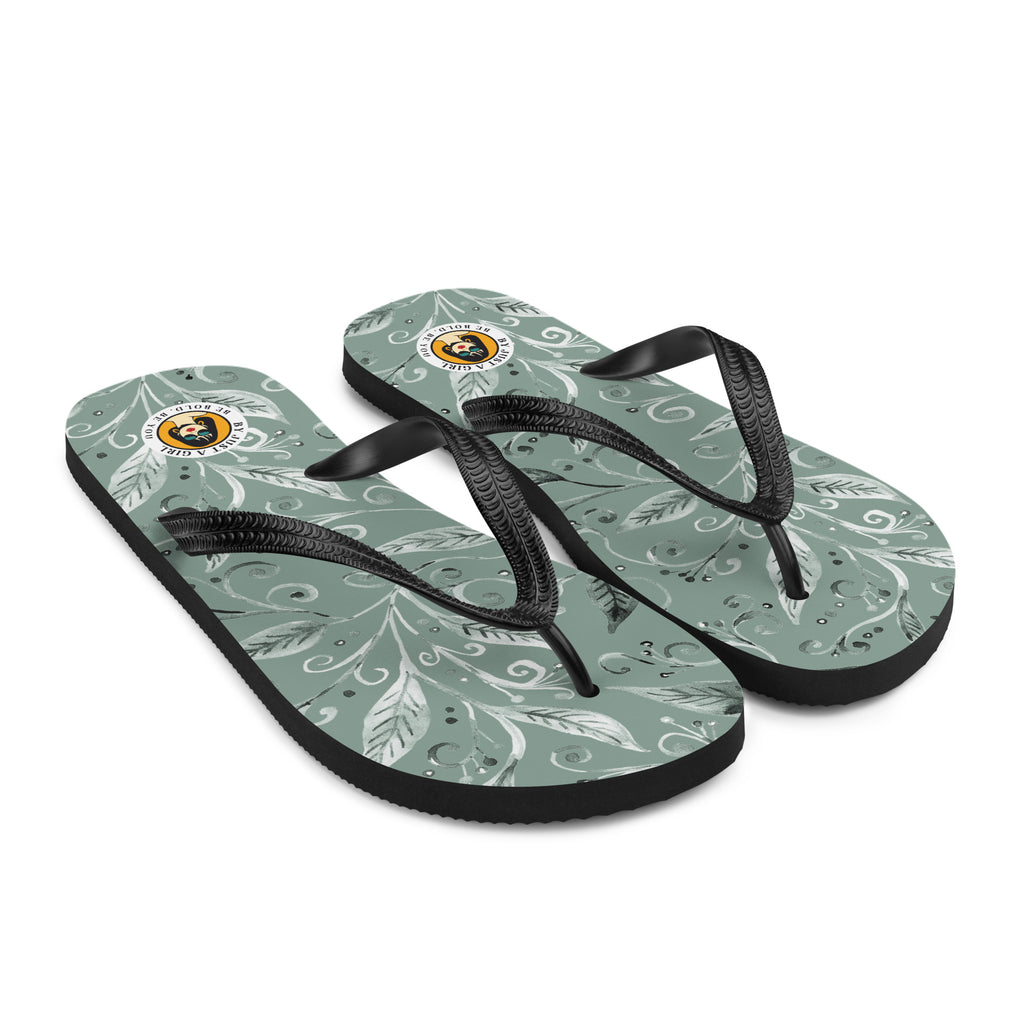 Ein Paar Flip-Flops mit einem Obermaterial in einem gedämpften Salbeigrün oder Grau-Grün. Das Muster besteht aus stilisierten weißen und hellgrauen Blättern, Ranken und kleinen Punkten, die organisch über die Oberfläche verteilt sind. Die Riemen sind schwarz und haben eine strukturierte Oberfläche. Auf der Ferse jedes Flip-Flops befindet sich ein rundes gelbes Emblem mit dem Text "BY JUST A GIRL" und "BE BOLD. BE YOU" sowie dem stilisierten Kopf einer Frau mit Bob-Frisur und Sonnenbrille.