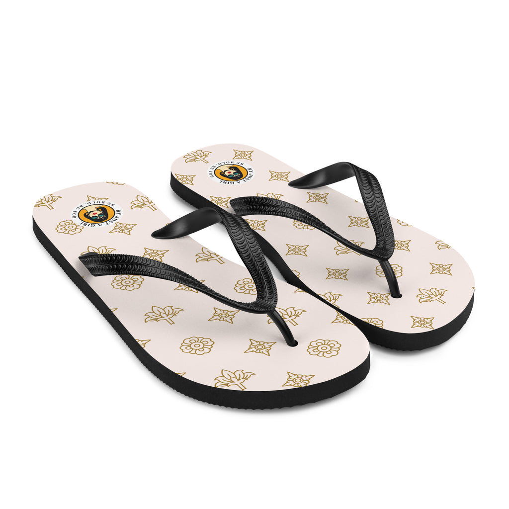 Ein Paar Flip-Flops mit einem hellbeigen oder cremefarbenen Obermaterial, das mit einem wiederkehrenden Muster aus stilisierten goldenen Lotusblüten und anderen floralen oder blattartigen Ornamenten bedeckt ist. Die Riemen sind schwarz und haben eine strukturierte Oberfläche. Auf der Ferse jedes Flip-Flops befindet sich ein rundes gelbes Emblem mit dem Text "BY JUST A GIRL" und "BE BOLD. BE YOU" sowie dem stilisierten Kopf einer Frau mit Bob-Frisur und Sonnenbrille.
