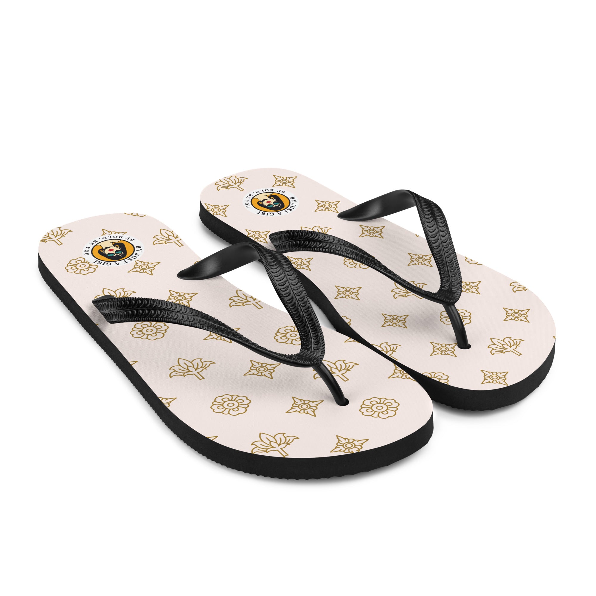 Ein Paar Flip-Flops mit einem hellbeigen oder cremefarbenen Obermaterial, das mit einem wiederkehrenden Muster aus stilisierten goldenen Lotusblüten und anderen floralen oder blattartigen Ornamenten bedeckt ist. Die Riemen sind schwarz und haben eine strukturierte Oberfläche. Auf der Ferse jedes Flip-Flops befindet sich ein rundes gelbes Emblem mit dem Text "BY JUST A GIRL" und "BE BOLD. BE YOU" sowie dem stilisierten Kopf einer Frau mit Bob-Frisur und Sonnenbrille.