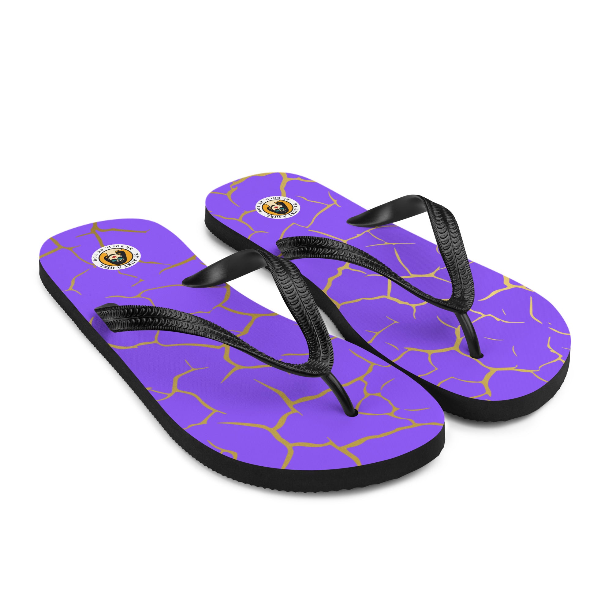 Ein Paar Flip-Flops mit einem Obermaterial in einem kräftigen Violett, das von einem unregelmäßigen, spinnennetzartigen Muster aus goldenen Linien durchzogen ist, die wie Risse aussehen. Die Riemen sind schwarz und haben eine strukturierte Oberfläche. Auf der Ferse jedes Flip-Flops befindet sich ein rundes gelbes Emblem mit dem Text "BY JUST A GIRL" und "BE BOLD. BE YOU" sowie dem stilisierten Kopf einer Frau mit Bob-Frisur und Sonnenbrille.